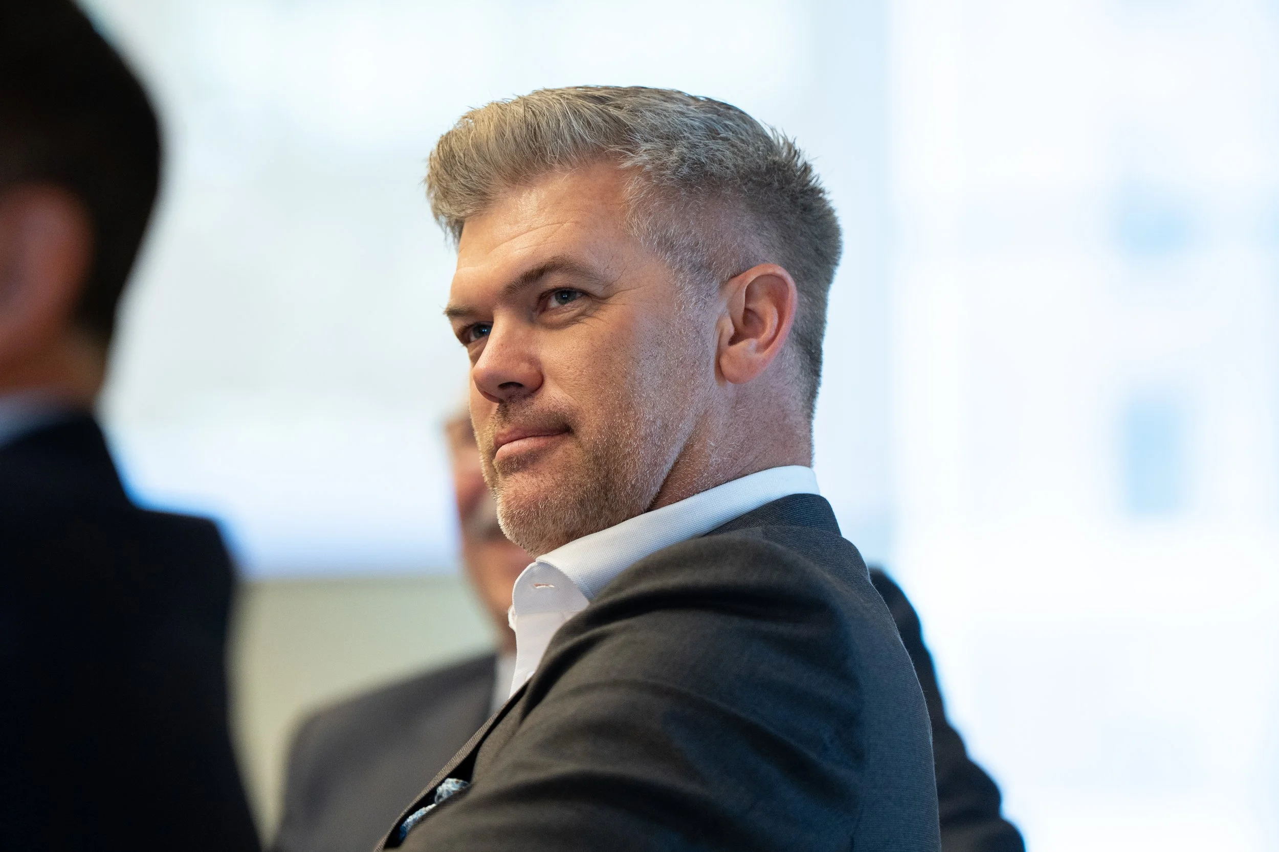 A man with short gray hair and a beard, wearing a black suit and white shirt, looking to the side during a professional event.
