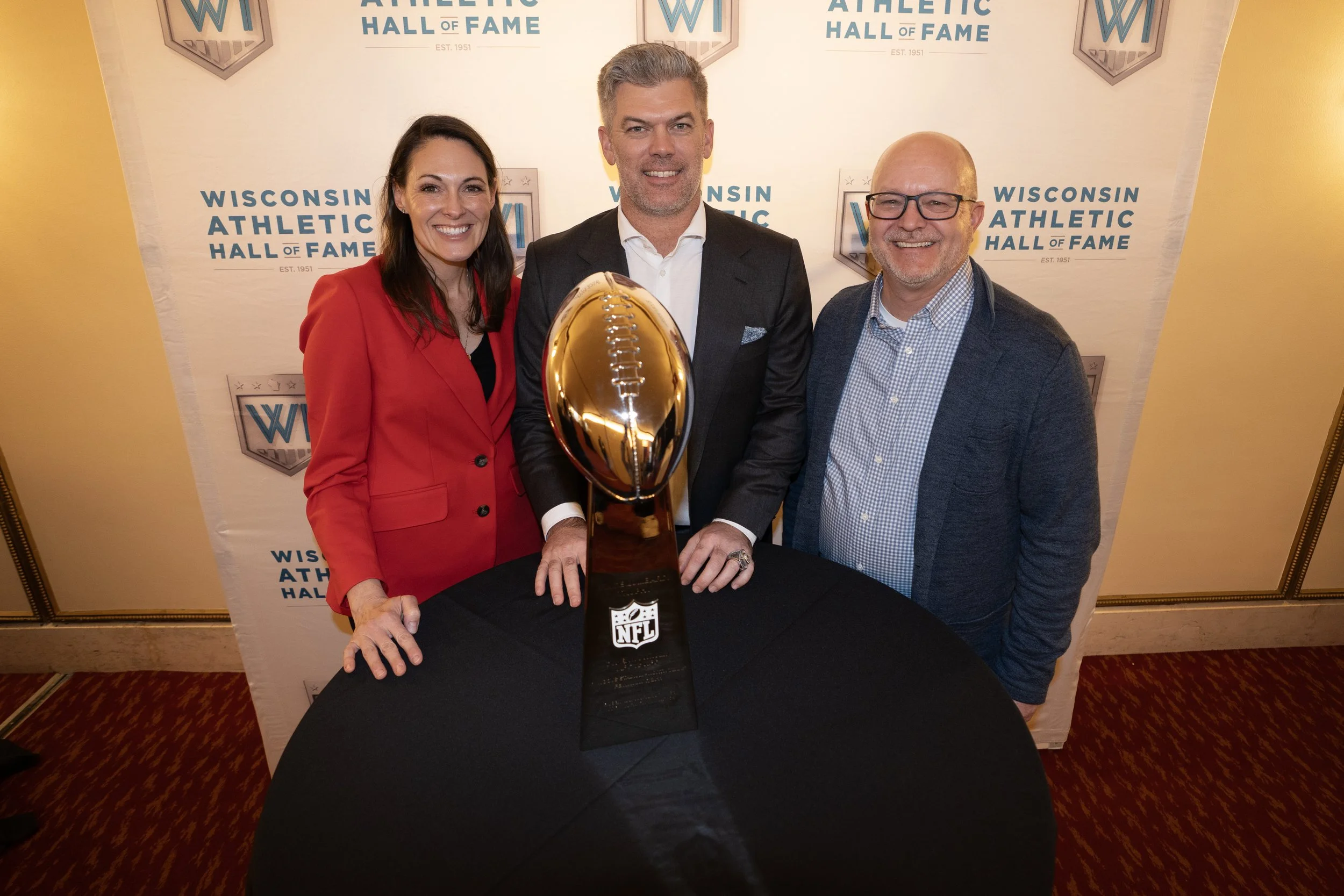 Three people, two men and a woman, standing behind a black table with a large, gold NFL trophy in the center. Background displays with the Wisconsin Athletic Hall of Fame logo and text.