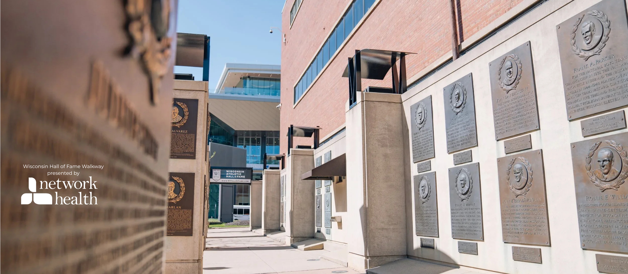 View of Wisconsin Hall of Fame plaques and statues on a brick and concrete wall at Wisconsin Hall of Fame Walkway outside a modern building with glass windows under a clear blue sky.