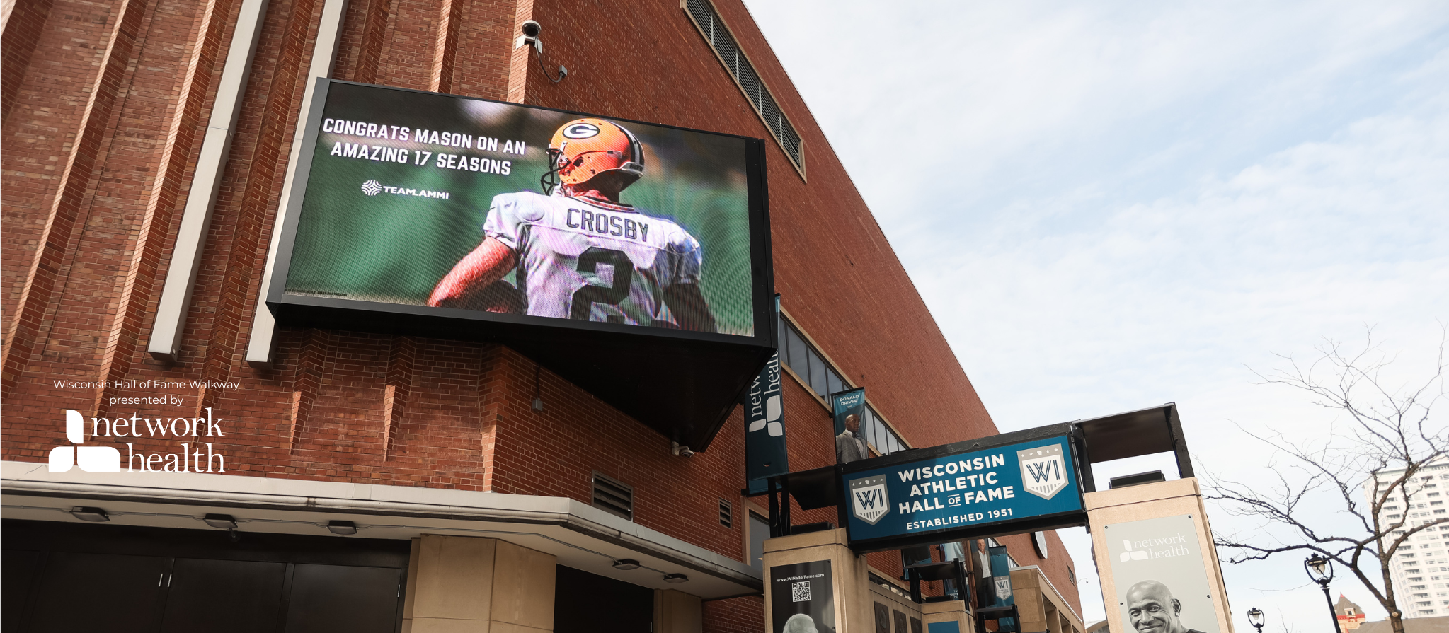 Outdoor view of Wisconsin Athletic Hall of Fame Walkway with digital video board advertisement showing a football player with the name 'Crosby' and number 2, celebrating a season, and a sign for Network Health.