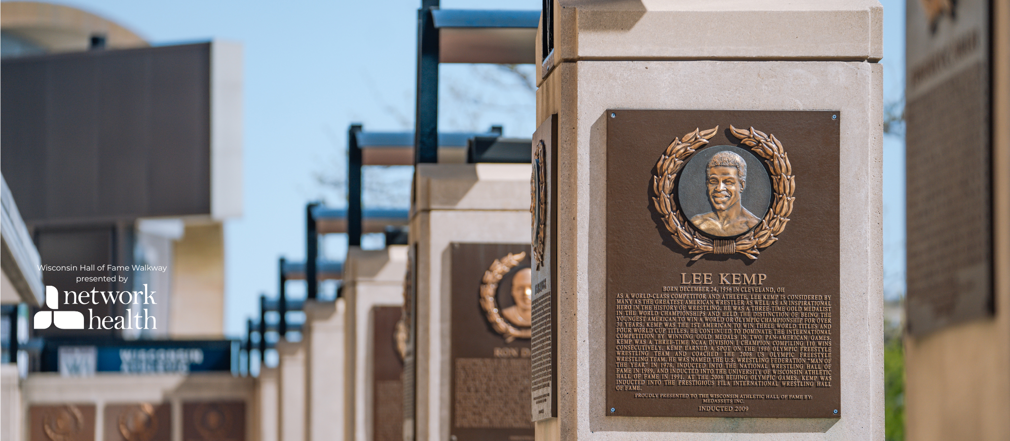 Close-up of memorial plaques on stone pillars at Wisconsin Hall of Fame Walkway, highlighting Lee Kemp's plaque with a portrait and biography.