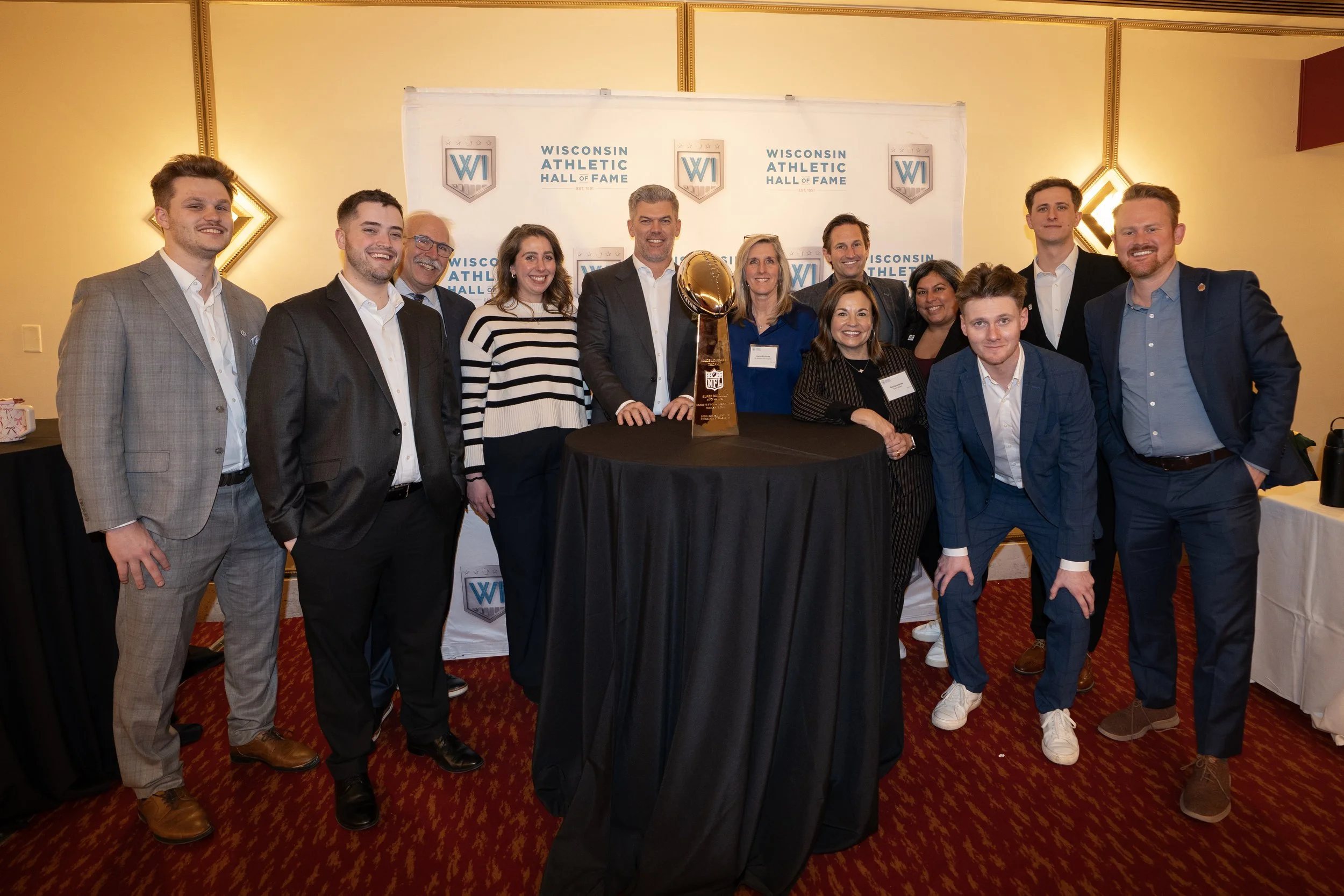A group of people at a Wisconsin Athletic Hall of Fame event, standing around a table with a large trophy, smiling and posing for a photo in a decorated room.