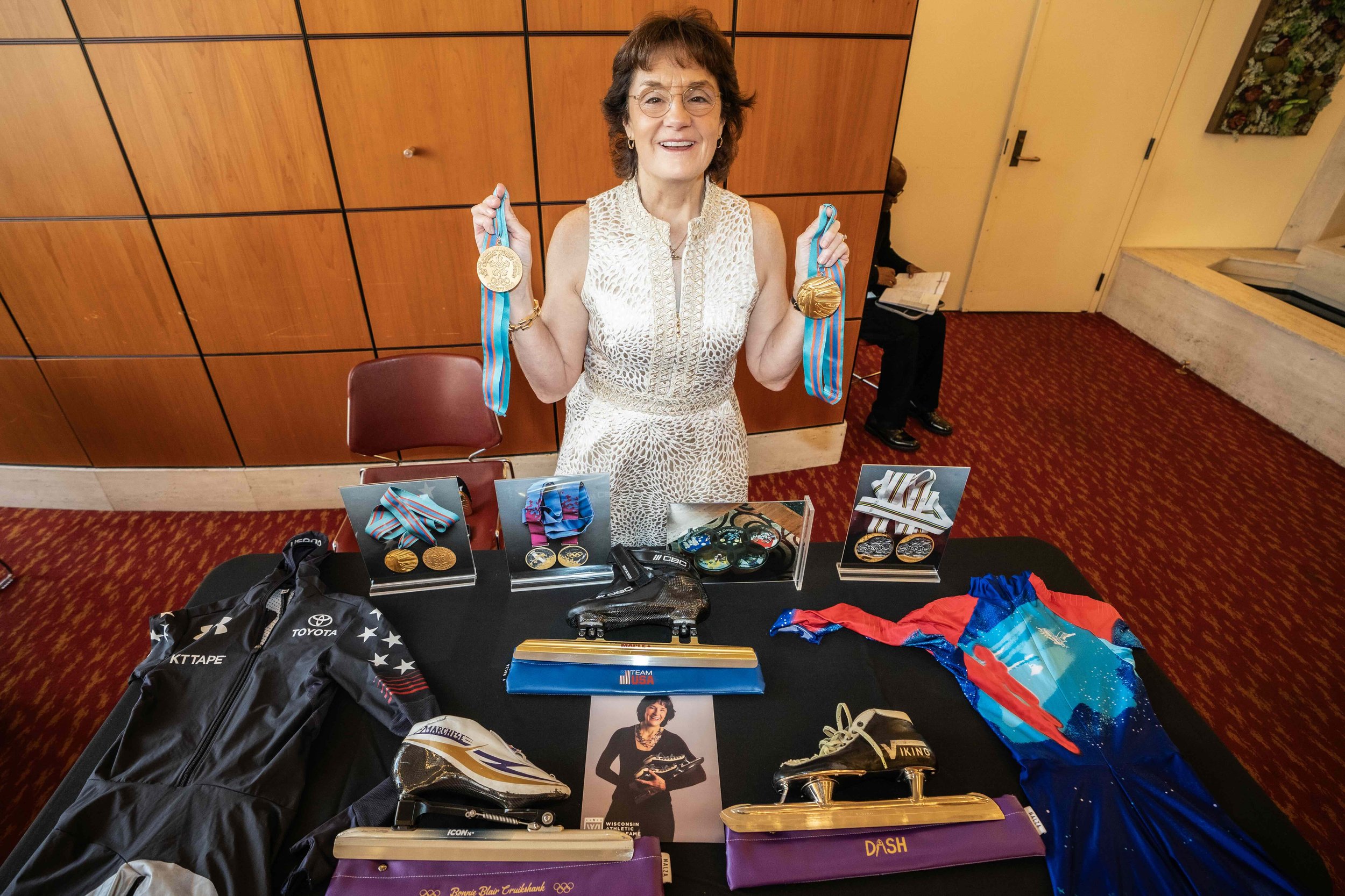 Smiling woman in sleeveless white dress holding two gold medals with blue ribbons at an awards table, which displays additional medals, medals in display cases, a racing jacket, skate shoes, and a photograph of the woman in a skating outfit.