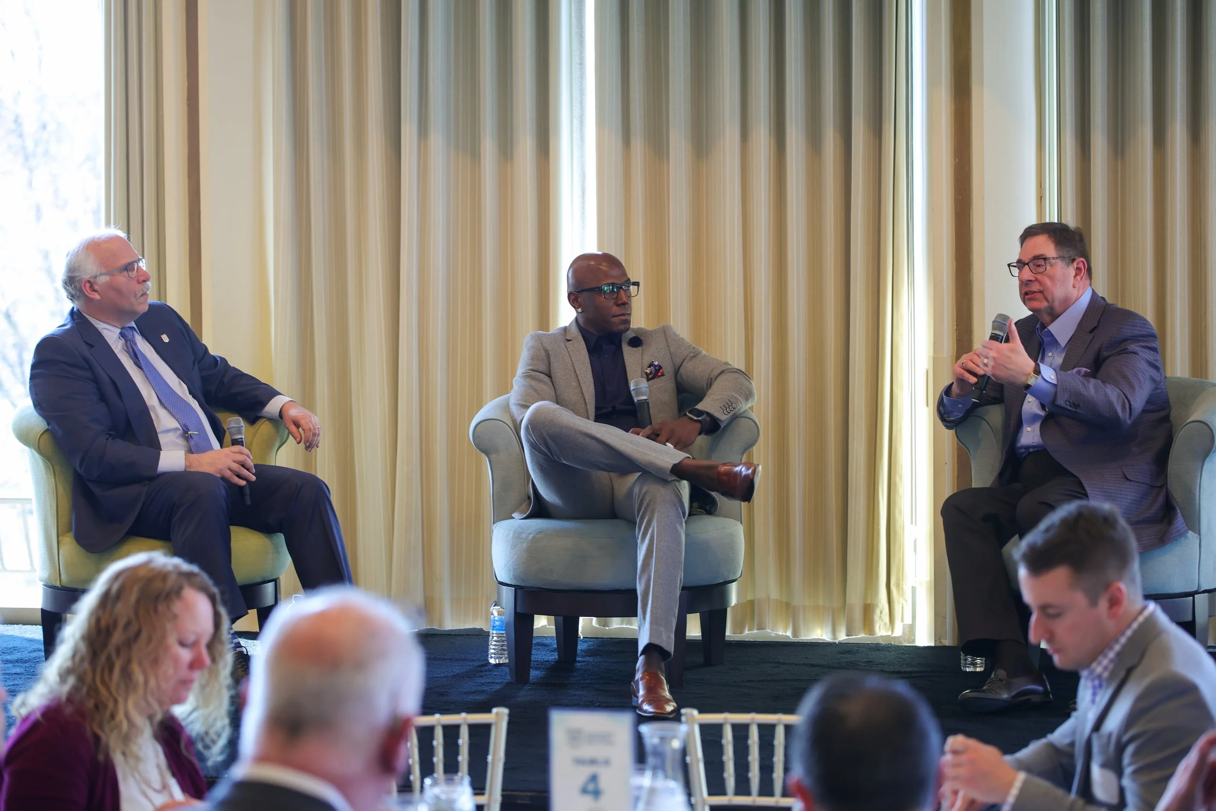 Three men sitting in chairs on a stage, engaging in a panel discussion at a conference or event. They are holding microphones and appear to be speaking to an audience.