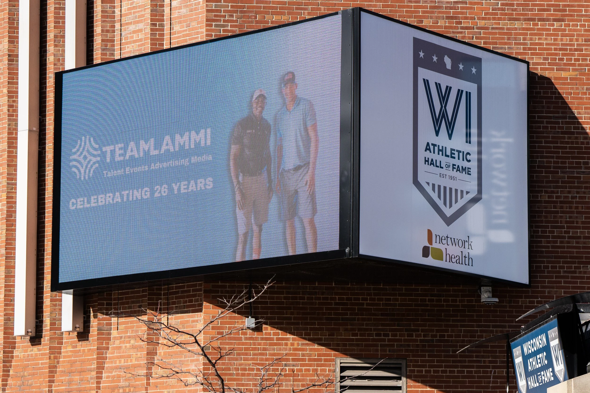 Digital billboard on brick building displays an image of two people standing together, with text celebrating 26 years of Teamlammi, a talent event advertising agency. Next to it is the Wisconsin Athletic Hall of Fame logo and a Network Health logo.