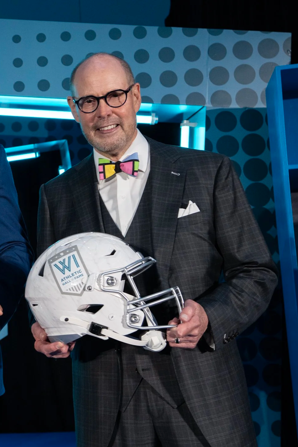 A man wearing glasses and a colorful bow tie holding a white football helmet with a logo that says WP Athletic Hall of Fame, standing in a room with blue and teal lighting and modern decor.
