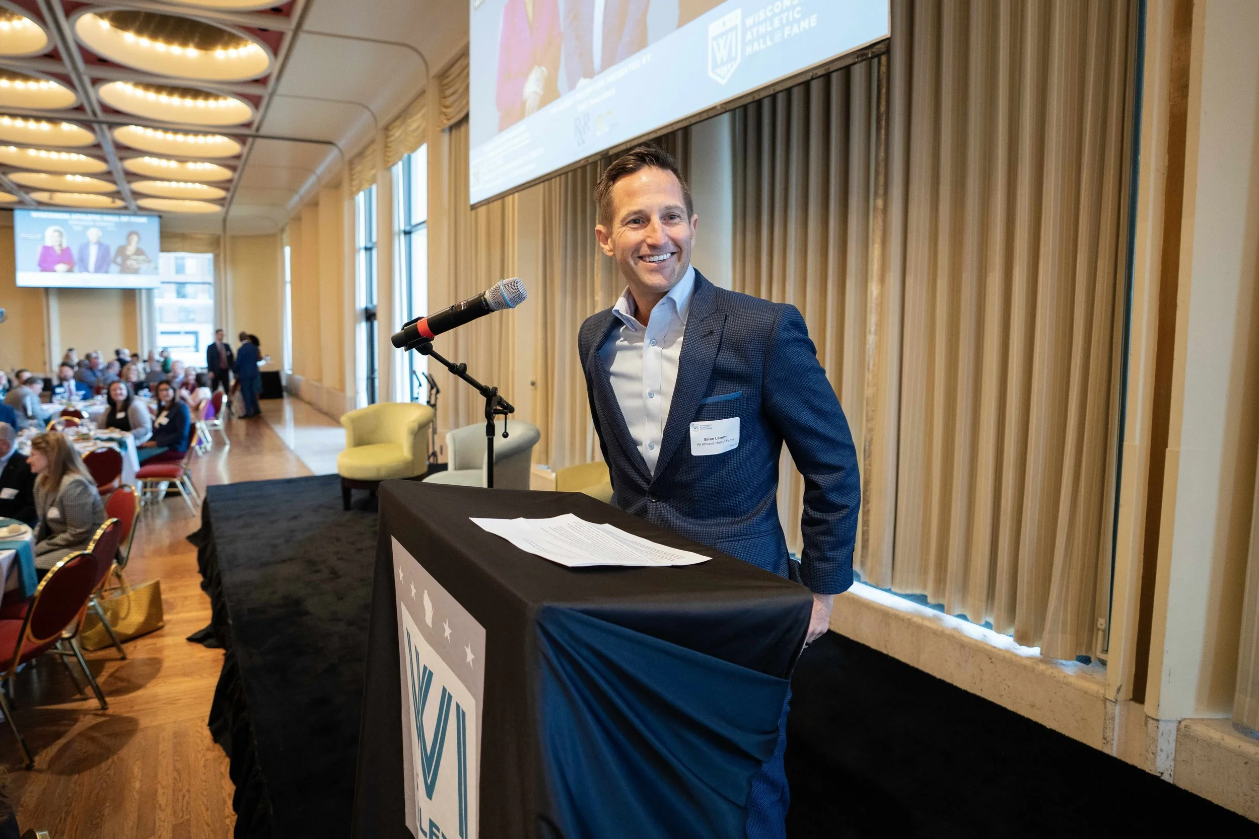 A man in a blue suit standing at a podium with a microphone, smiling, at a conference or event with people seated at tables and large screens in the background.