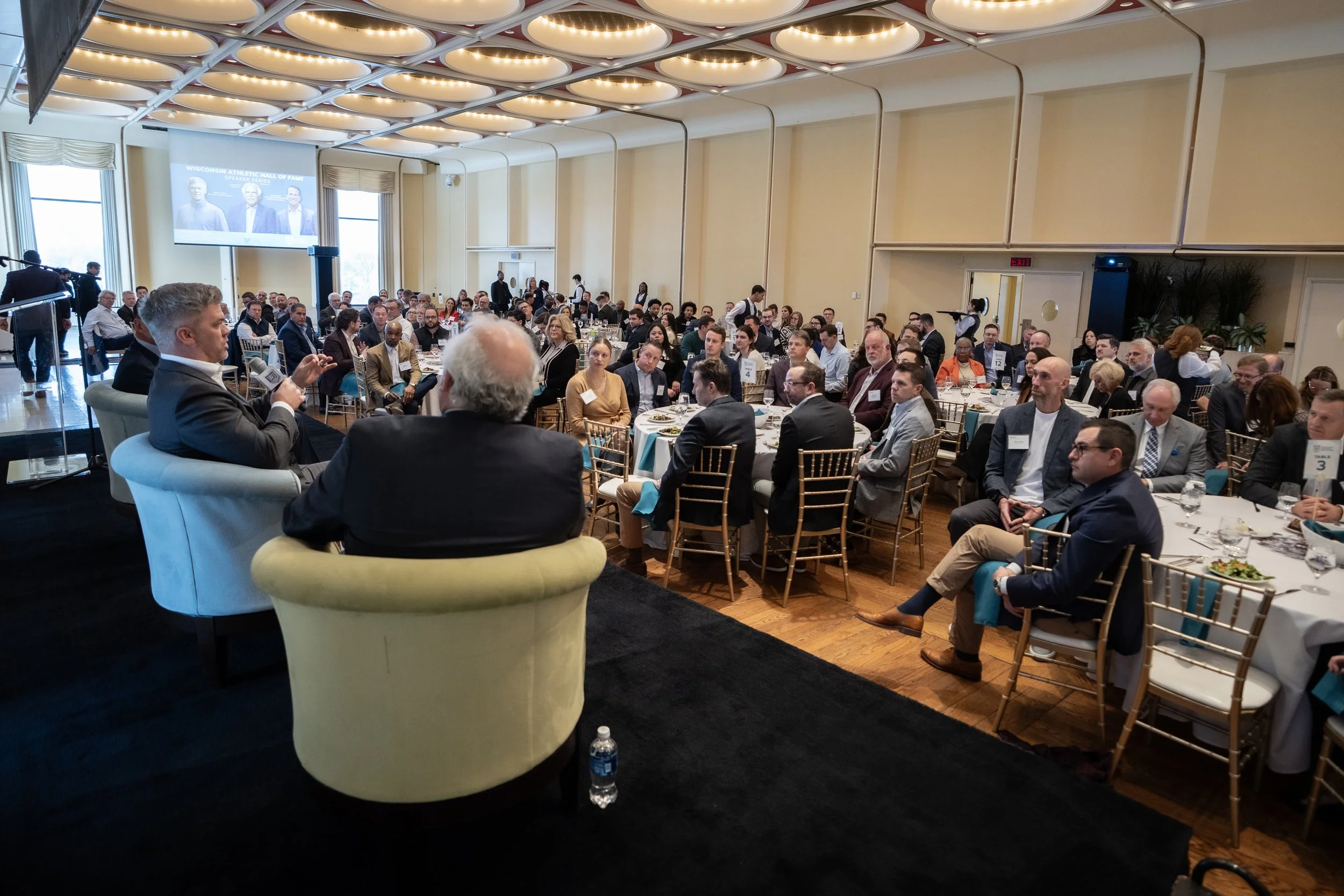 A large conference room filled with many people sitting at round tables, listening to a panel of speakers at the front. The room has bright lighting, large windows, and a projection screen displaying images and text.