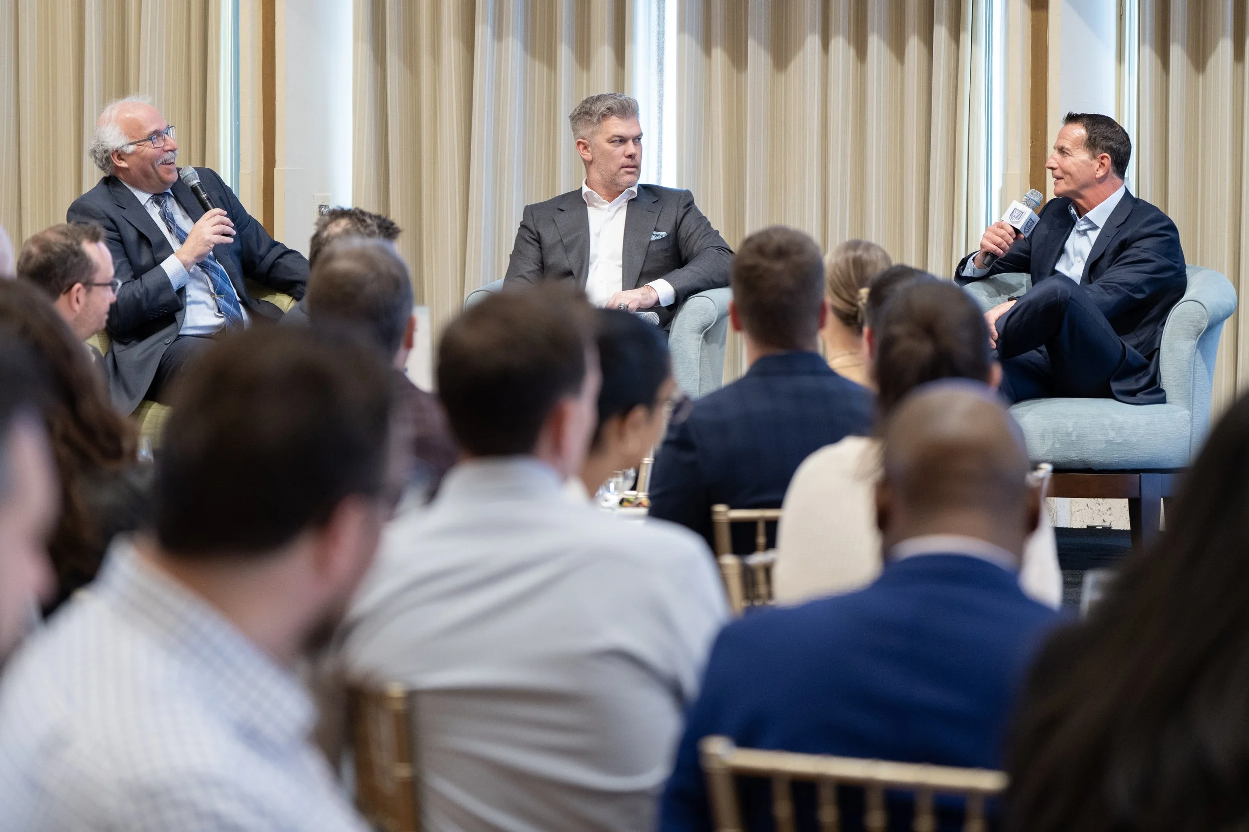 Three men are sitting on stage in conversation at a panel discussion, with an audience of diverse adults seated in front of them.