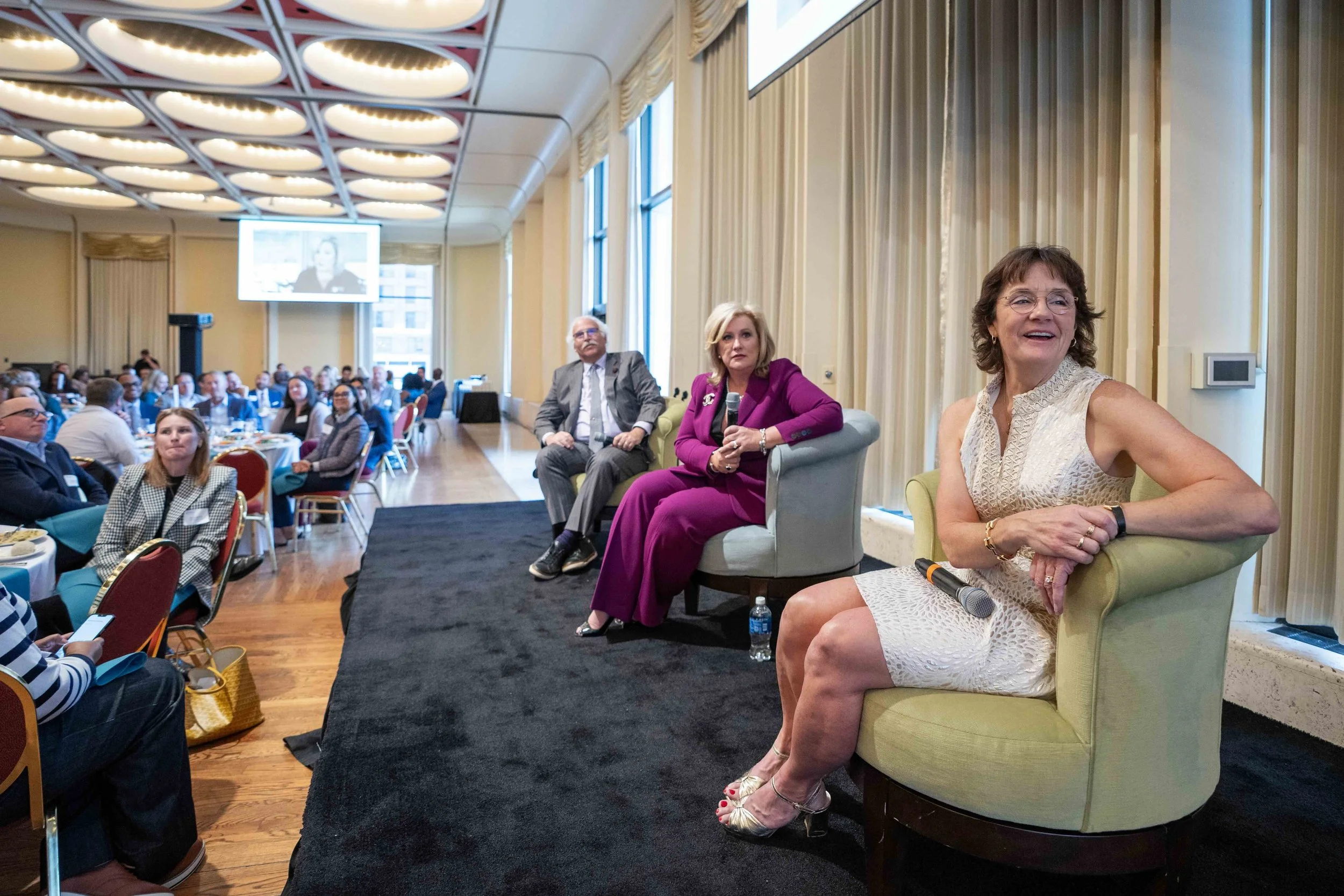 A panel discussion at a conference with three women seated on stage in front of an audience. The woman on the right is wearing a sleeveless cream dress, the woman in the middle is in a purple suit, and the woman on the left is in a light-colored blaz