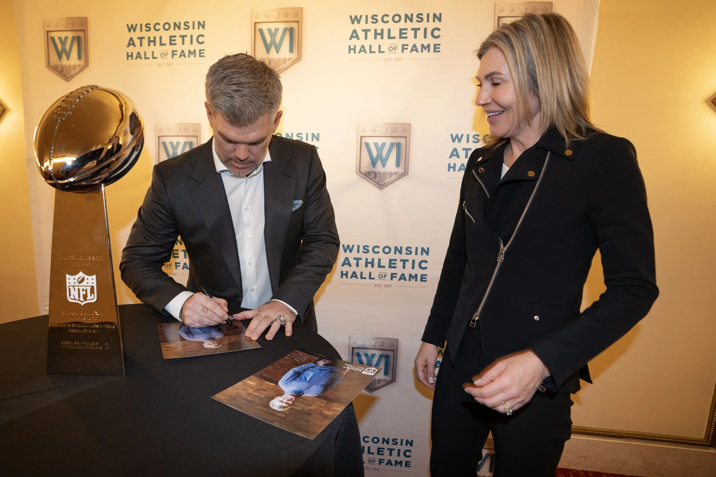A man in a suit is signing a photograph at a Wisconsin Athletic Hall of Fame event, with a woman in black standing next to him. A Super Bowl NFL trophy is on the table, in front of a backdrop with the Wisconsin Athletic Hall of Fame logo.