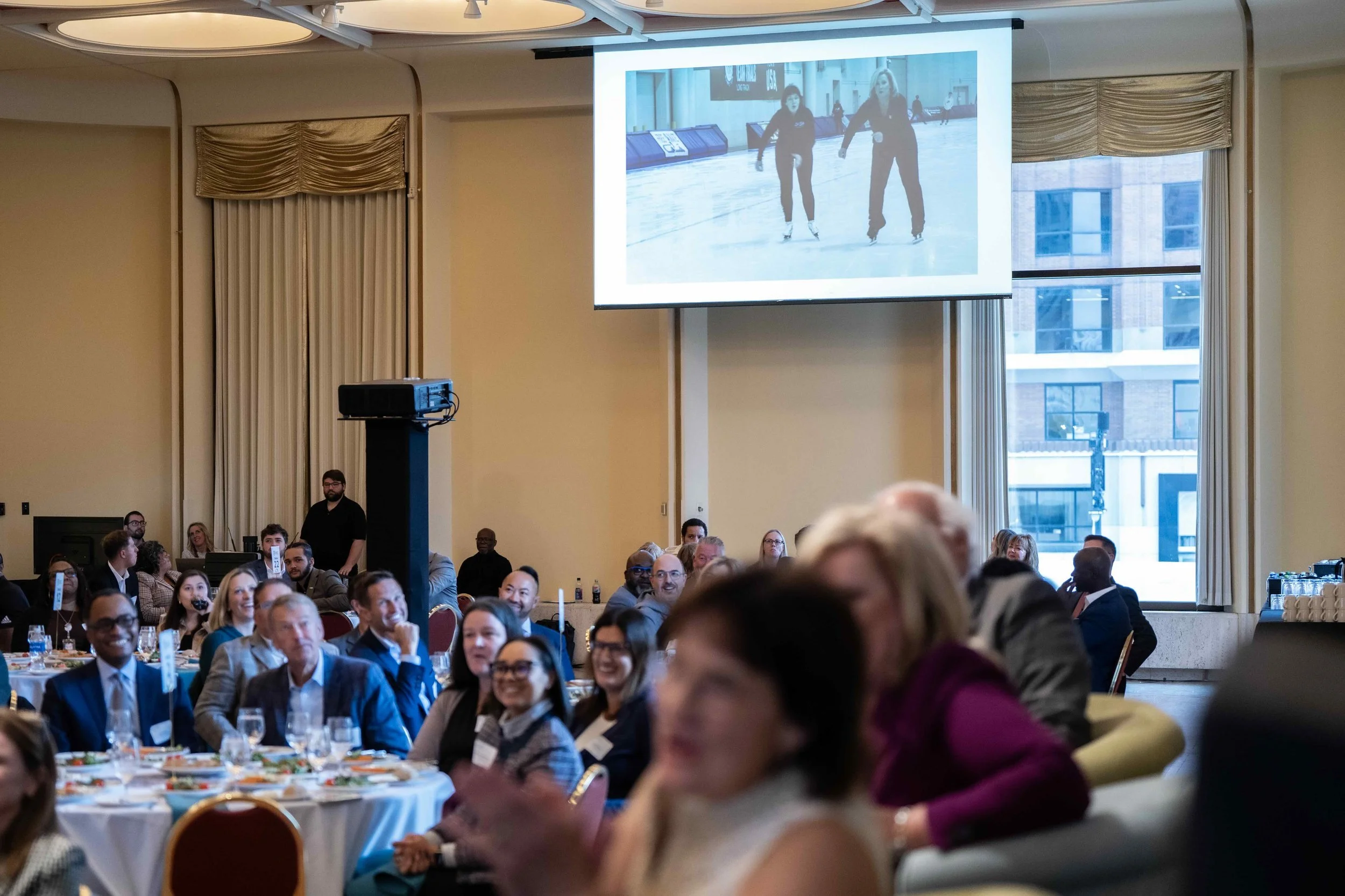 People attending a conference or event seated at round tables, watching a presentation displayed on a large screen, with some smiling and engaging. The room has large windows, curtains, and a high ceiling.
