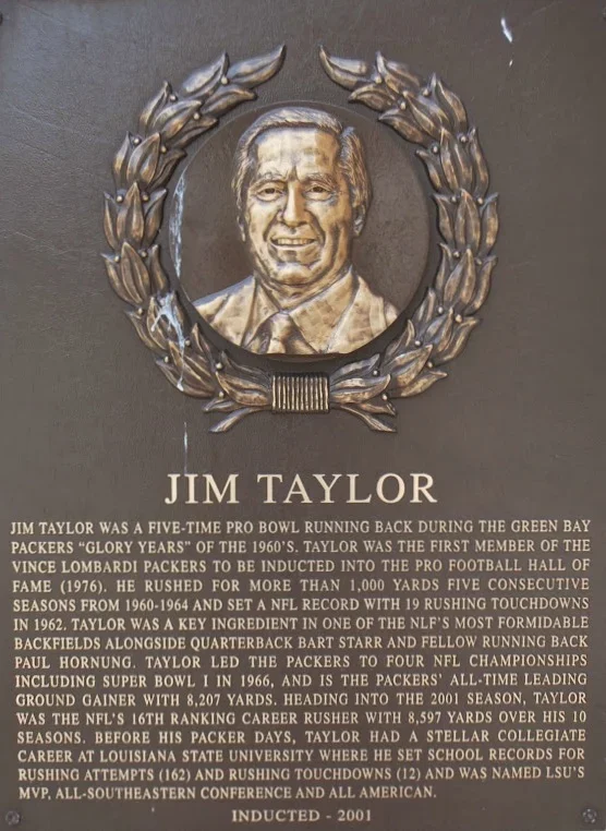 Plaque with a portrait of Jim Taylor, a former professional football player, surrounded by a decorative wreath. The plaque details his achievements, including five Pro Bowl selections, NFL rushing record, and college accomplishments at LSU.
