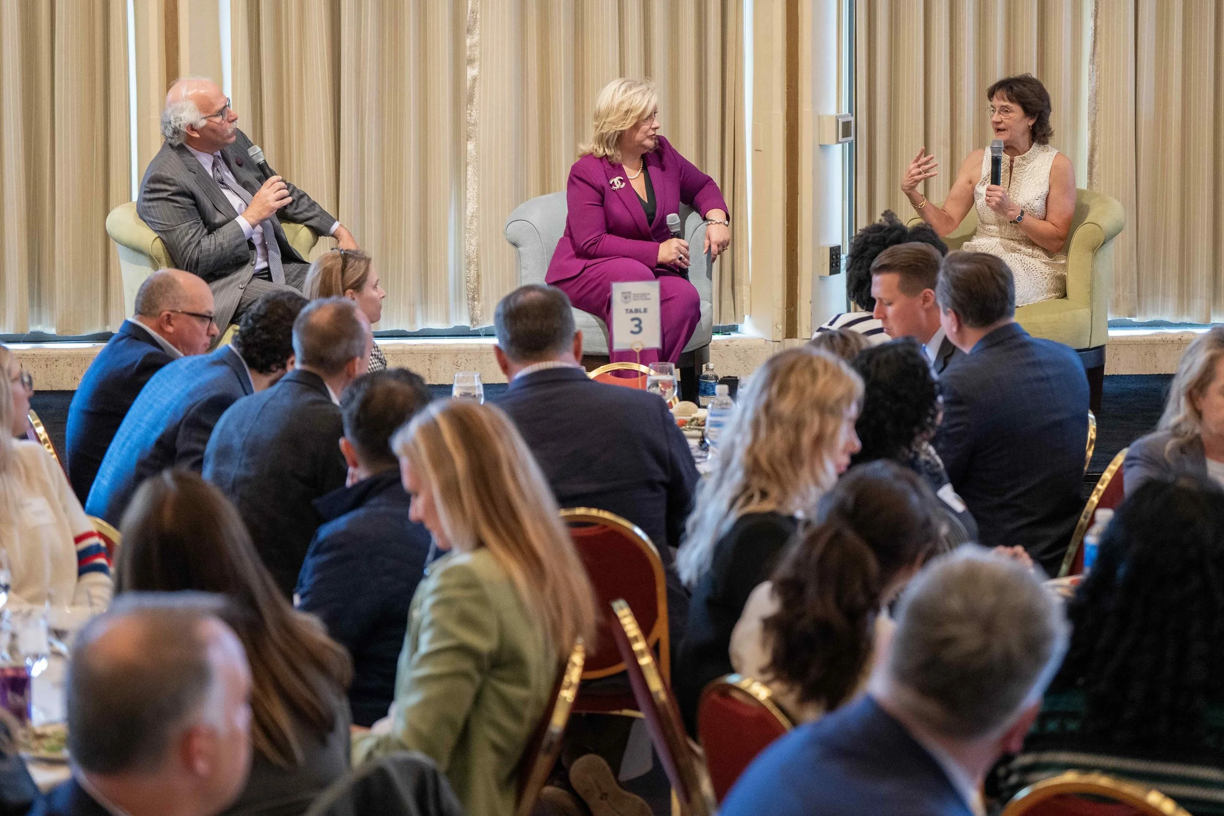 A panel discussion at a conference with three women on stage, two speaking into microphones while seated in armchairs, and a seated audience in front of them.