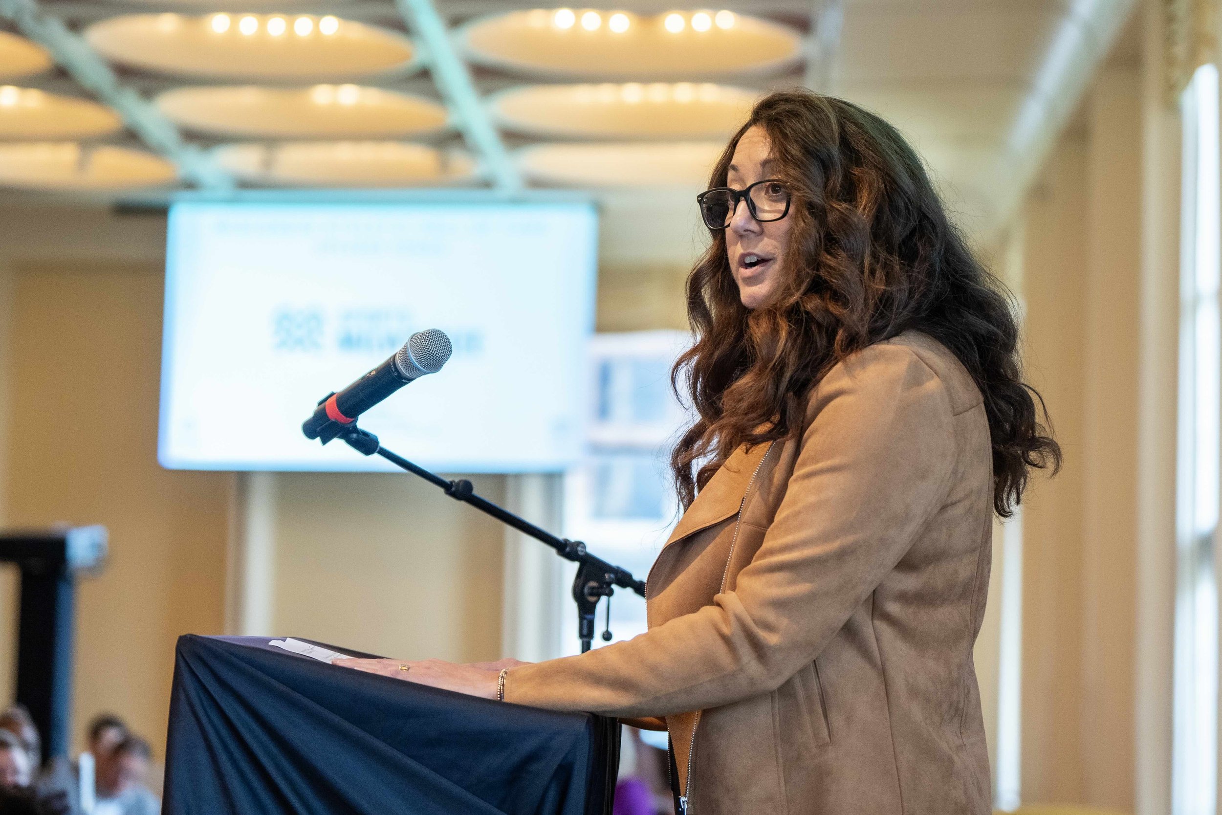 A woman with curly hair, glasses, and a brown jacket is speaking into a microphone at a podium during a conference or event, with a blurred screen and audience in the background.