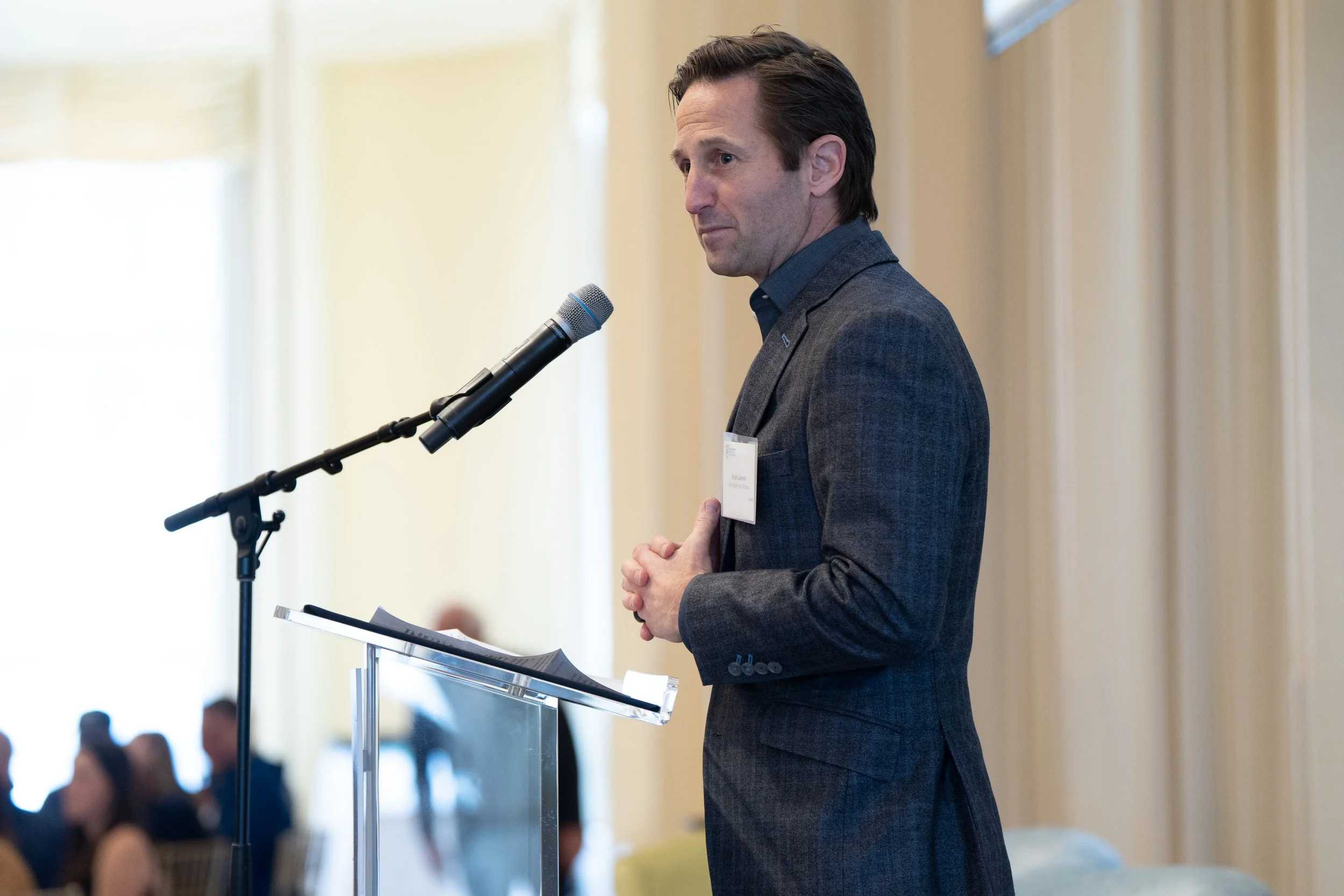 A man in a dark gray suit standing at a podium with a microphone, speaking or listening during a conference or event, with a blurred audience in the background.