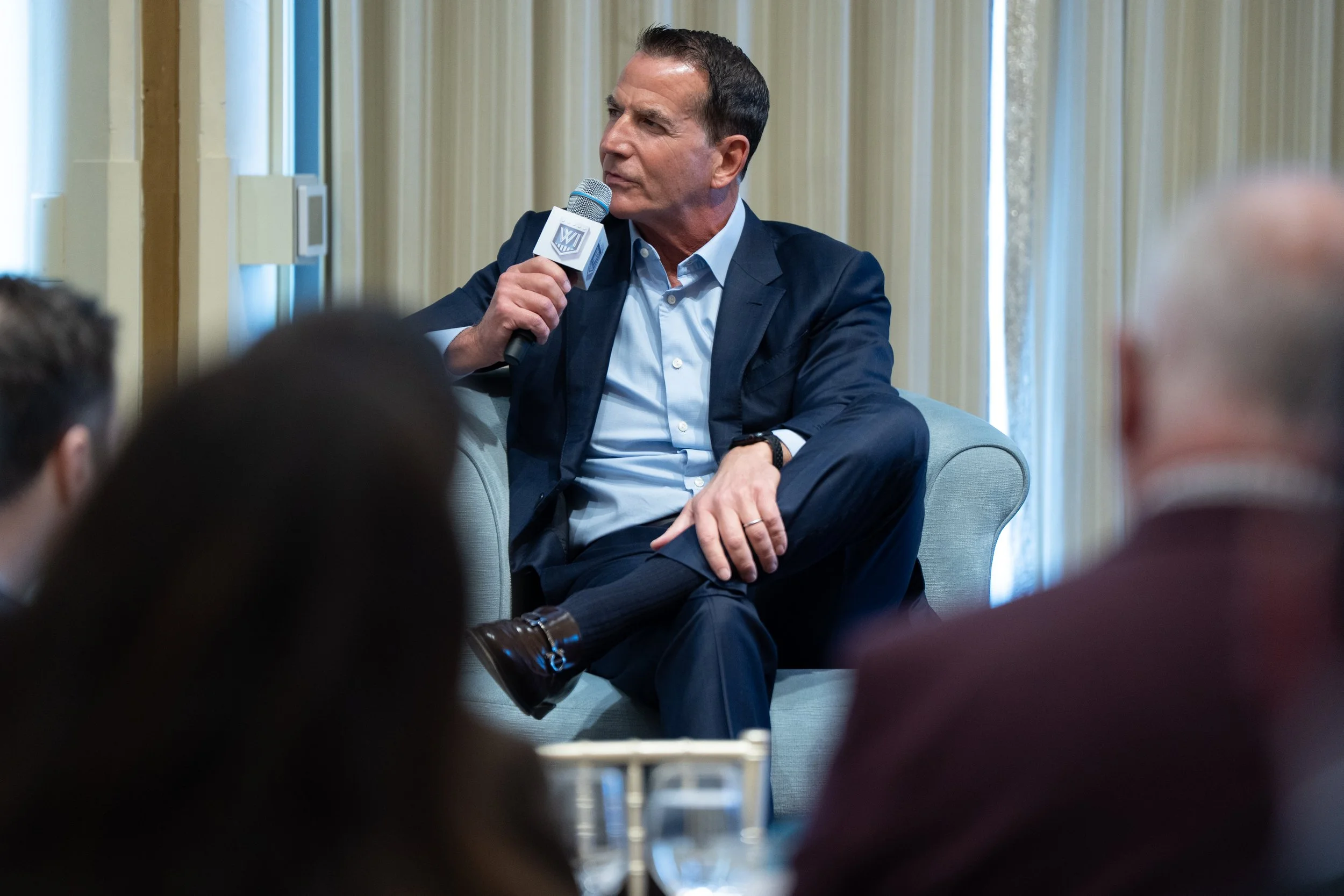 A man in a suit sitting in an armchair, holding a microphone, speaking at a conference or panel discussion with an audience.