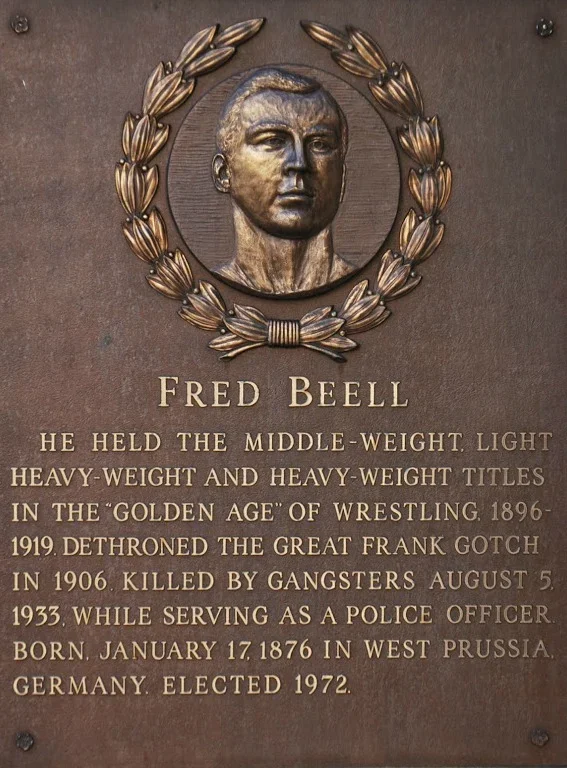 A commemorative plaque featuring a metal relief portrait of Fred Beell, surrounded by a wreath. The plaque contains text about Fred Beell's life, achievements in wrestling, and service as a police officer, including his birth date and place, and political career.