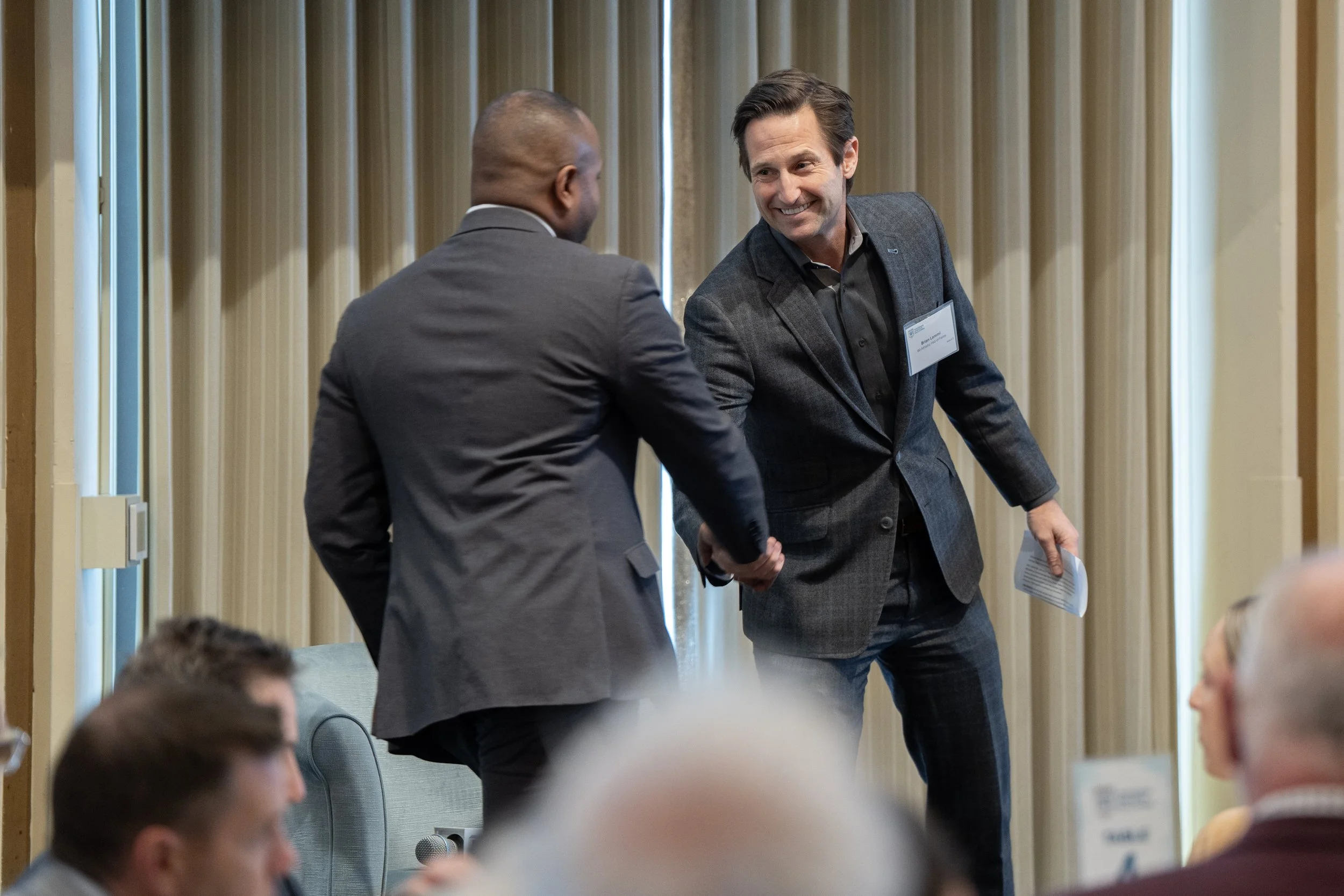 Two men in suits shaking hands at a professional event or conference, with a seated audience in the foreground.