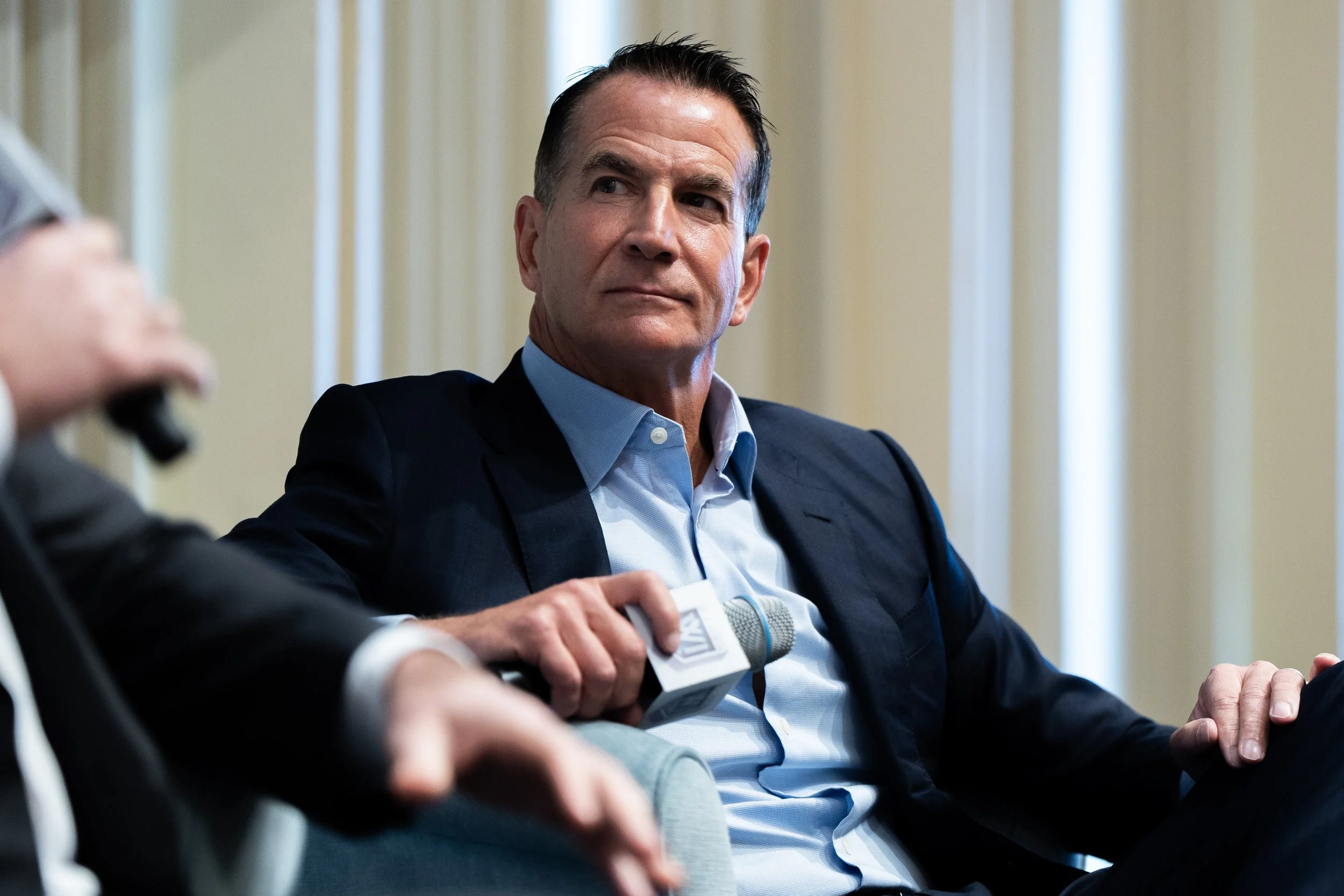 Man in a suit participating in a panel discussion, holding a microphone, with a serious expression, in a formal indoor setting.