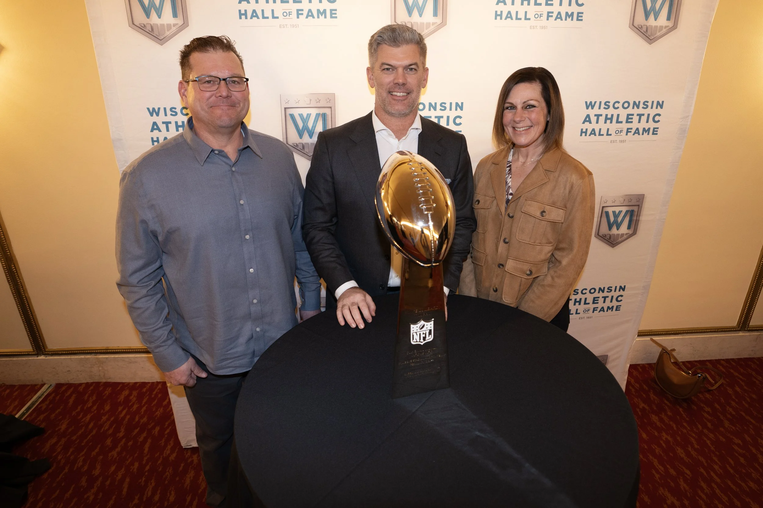 Three people standing behind a table with a large NFL Super Bowl trophy, smiling, at the Wisconsin Athletic Hall of Fame event. Two men and one woman, all dressed in semi-formal attire, with a backdrop featuring the event's logo.