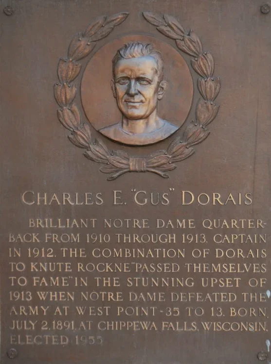 Bronze plaque with a relief portrait of Charles E. "Gus" Dorais, a football player, surrounded by a laurel wreath. The plaque contains an inscription honoring his achievements as a Notre Dame quarterback from 1910 to 1913, his role as captain in 1912, and mentions his military service and election in 1955.