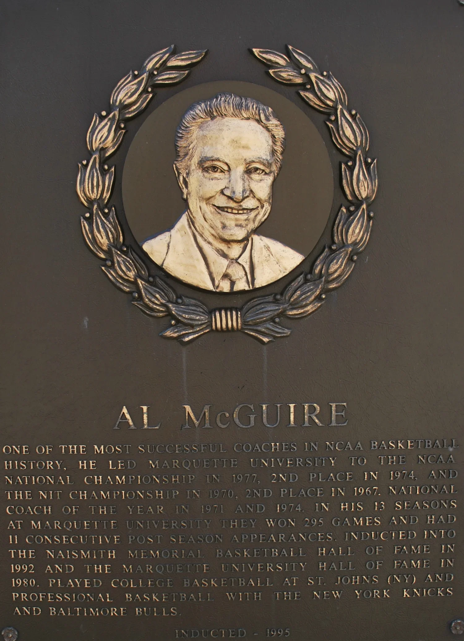 A commemorative plaque engraved with a portrait of Al McGuire and an inscription about his achievements as a basketball coach, including his work at Marquette University and recognition in the Naismith Memorial Basketball Hall of Fame.
