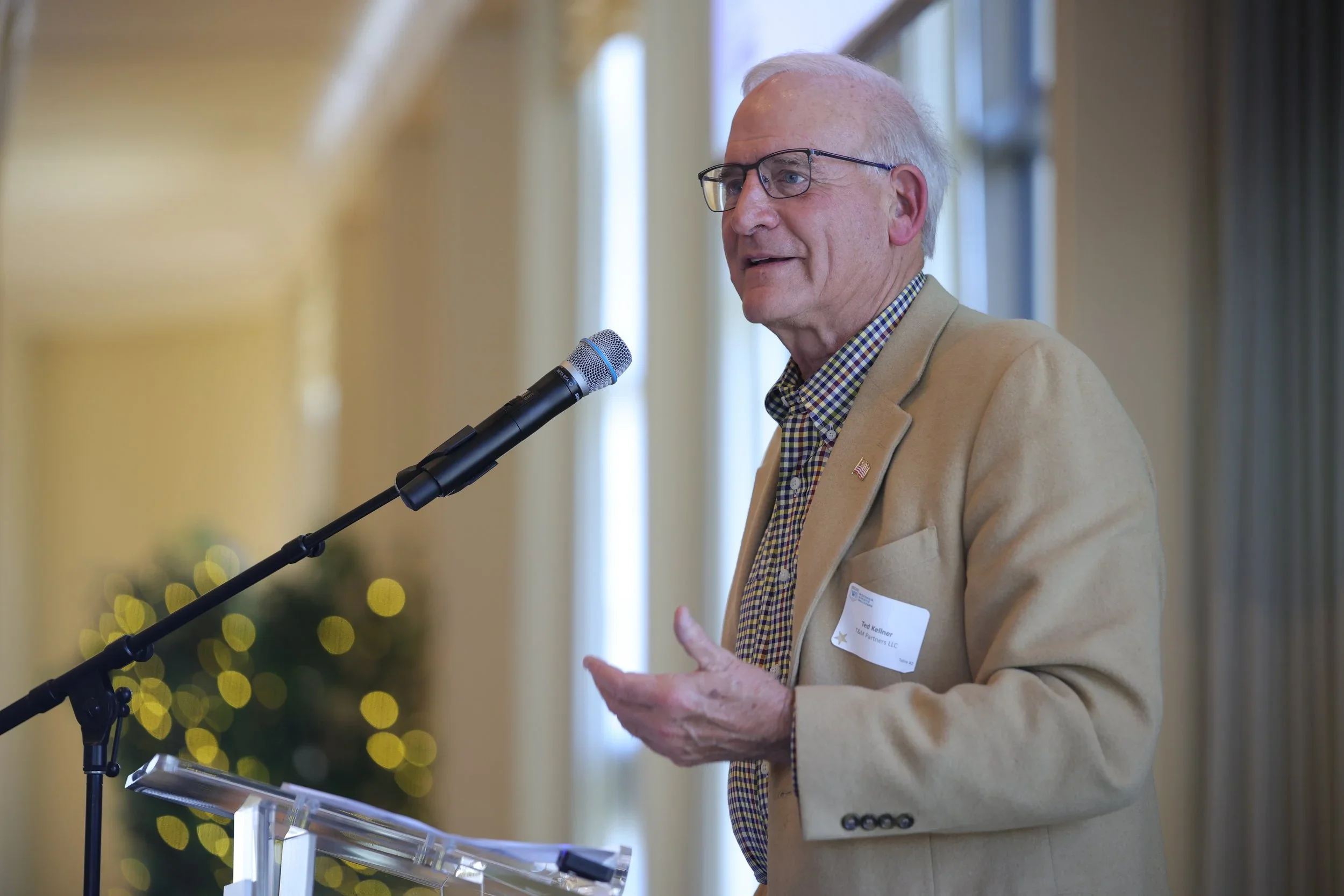 An elderly man with glasses, wearing a tan blazer and checkered shirt, is speaking into a microphone at a podium indoors. There is a blurred background with holiday lights and windows.
