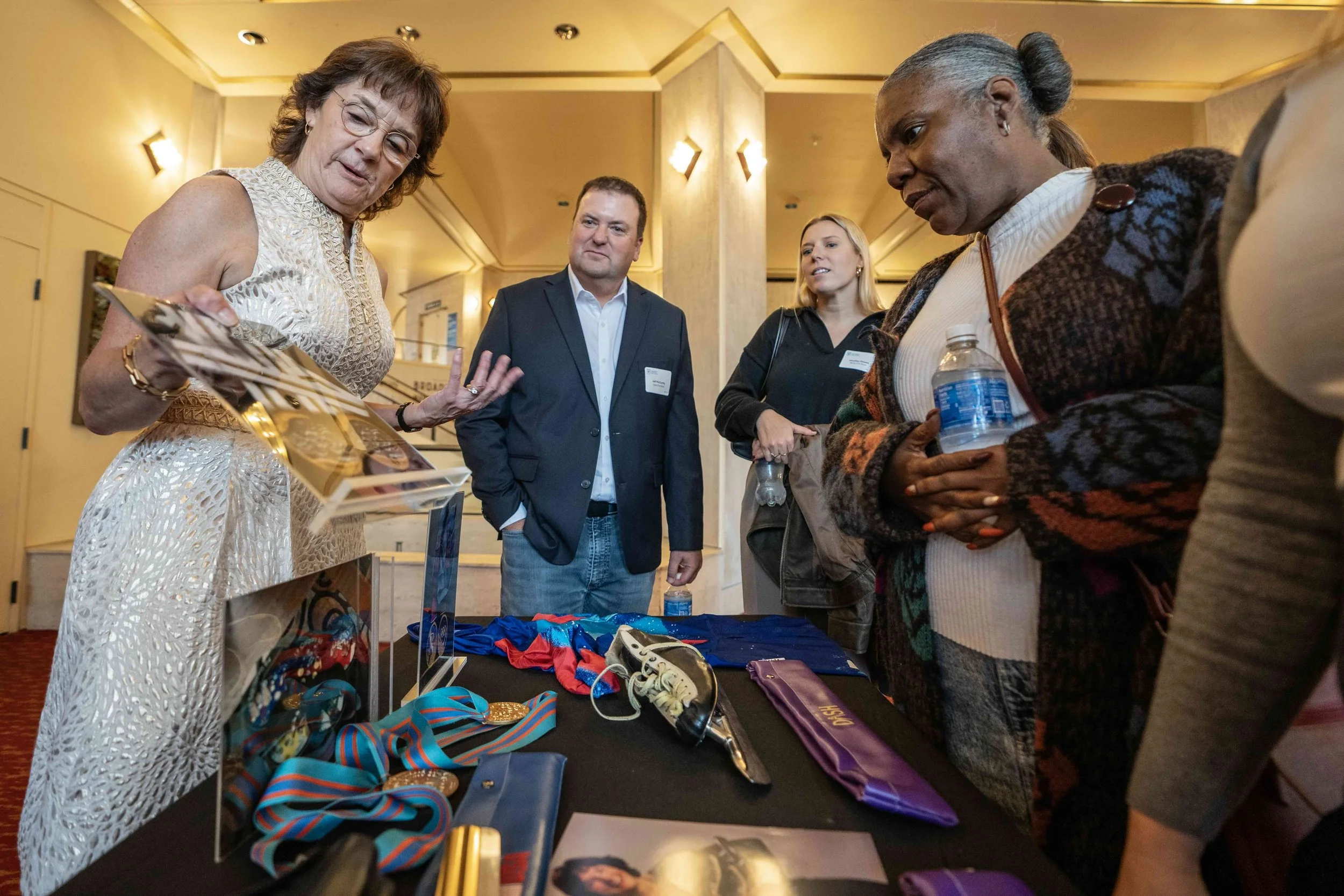A woman showing various medals and ribbons on a table to a group of people at an indoor event.
