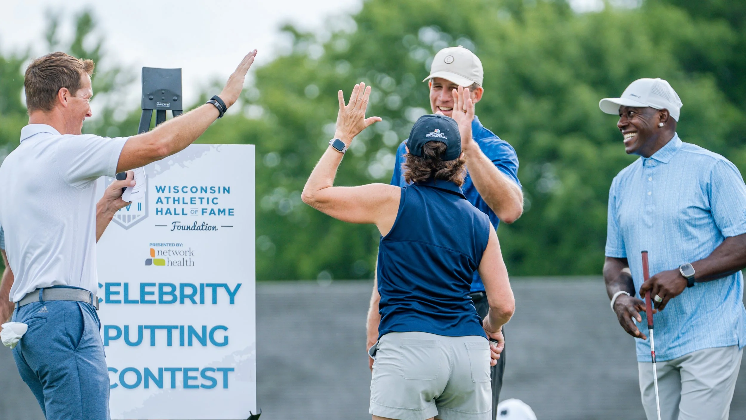 Group of people at a golf event, smiling and celebrating, with one woman giving a high five to a man, and others laughing and enjoying the outdoor setting.