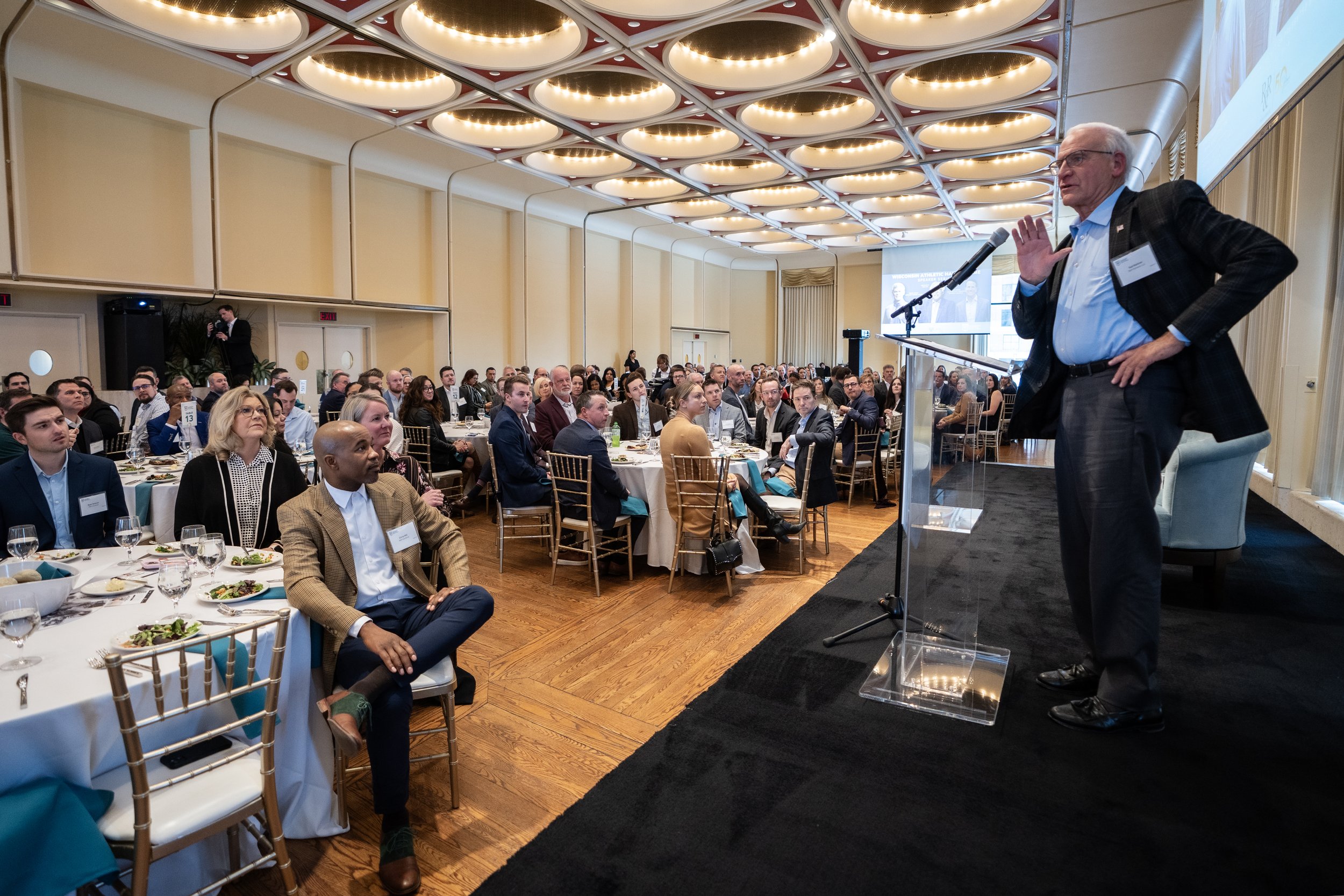 A man speaking at a podium during a conference or banquet, with an audience of men and women seated at round tables with food and drinks, in an elegant banquet hall with a high ceiling and decorative lighting.