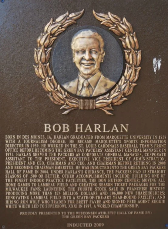 Plaque with a portrait of Bob Harlan surrounded by a decorative laurel wreath. The plaque provides a biography of Bob Harlan, highlighting his education at Marquette University, tenure with the Green Bay Packers, and notable achievements, including his induction into the Green Bay Packers Hall of Fame in 2009.