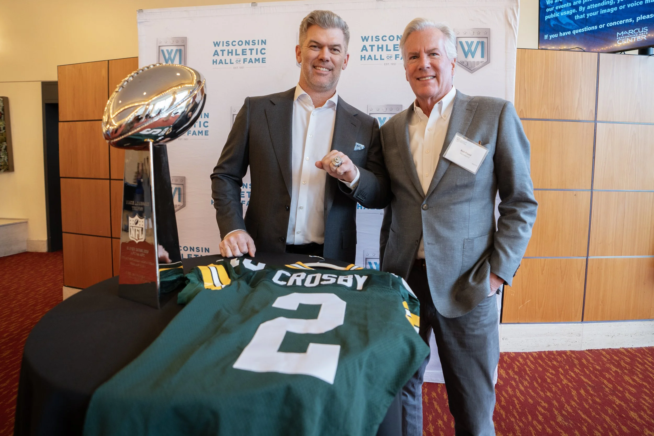 Two men in suits standing behind a table with a Green Bay Packers football jersey and a Lombardi trophy. The background features a banner with 'Wisconsin Athletic Hall of Fame' and the logos 'WI' and 'WISCONSIN ATHLETIC HALL OF FAME'. One man is show