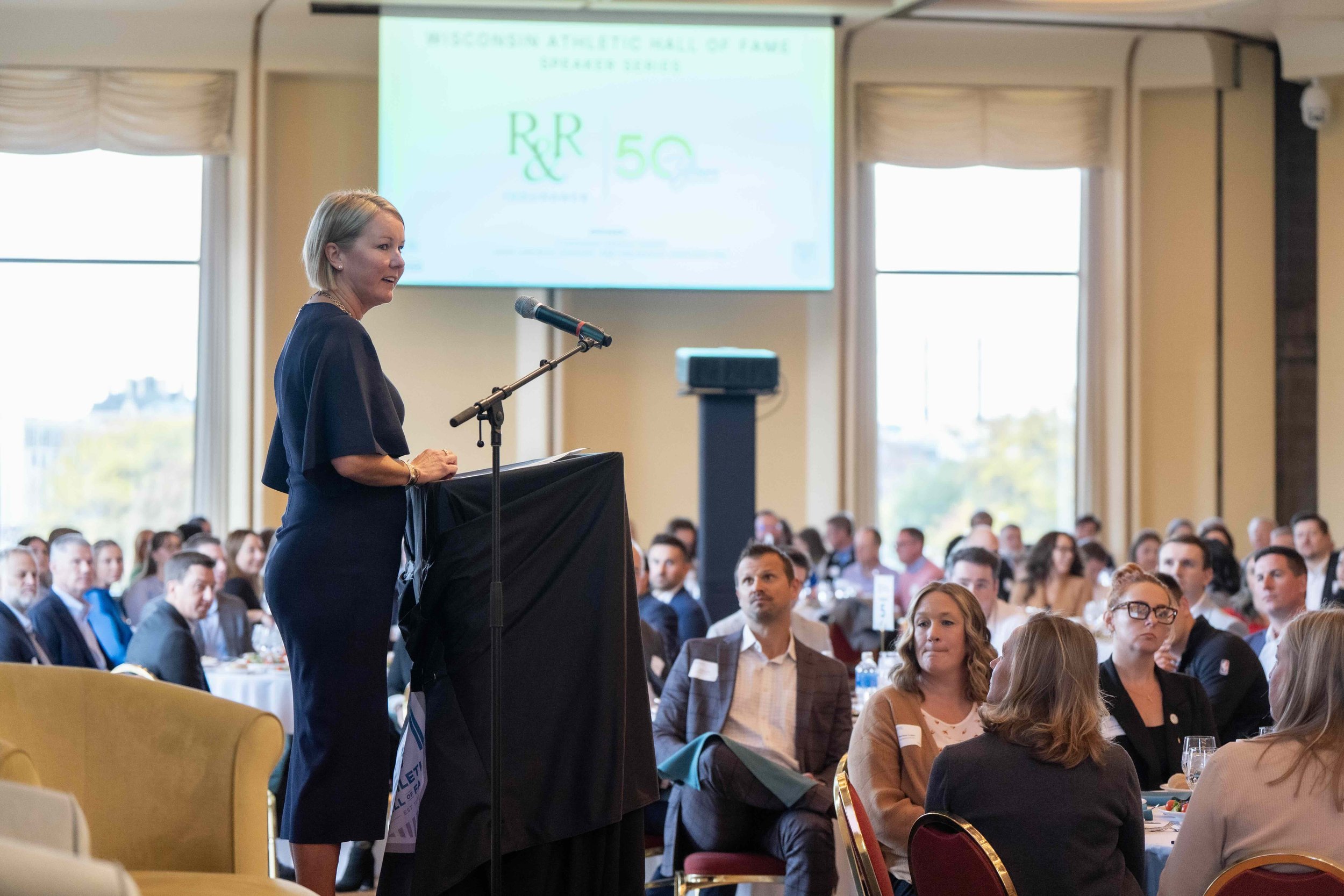 A woman giving a presentation at a conference, standing at a podium with a microphone, audience members seated at tables, large windows in the background, and a screen displaying a presentation slide.