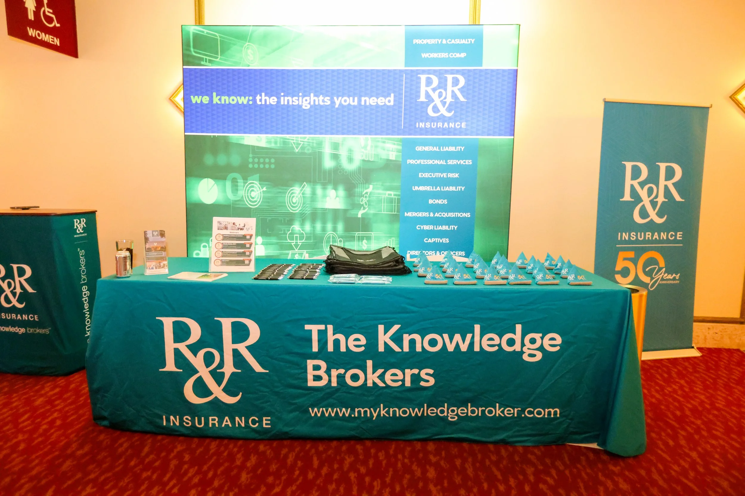 Trade show booth for R&R Insurance featuring a large banner with the text "we know: the insights you need" and company details, a table with branded promotional items including pens and brochures, a teal tablecloth with R&R Insurance branding, a teal