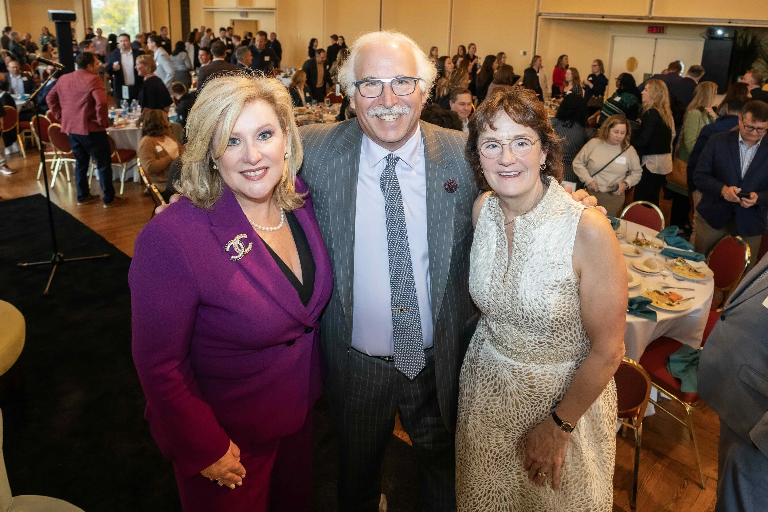 Three people smiling at a formal event, standing close together inside a decorated banquet hall with tables and other guests in the background.