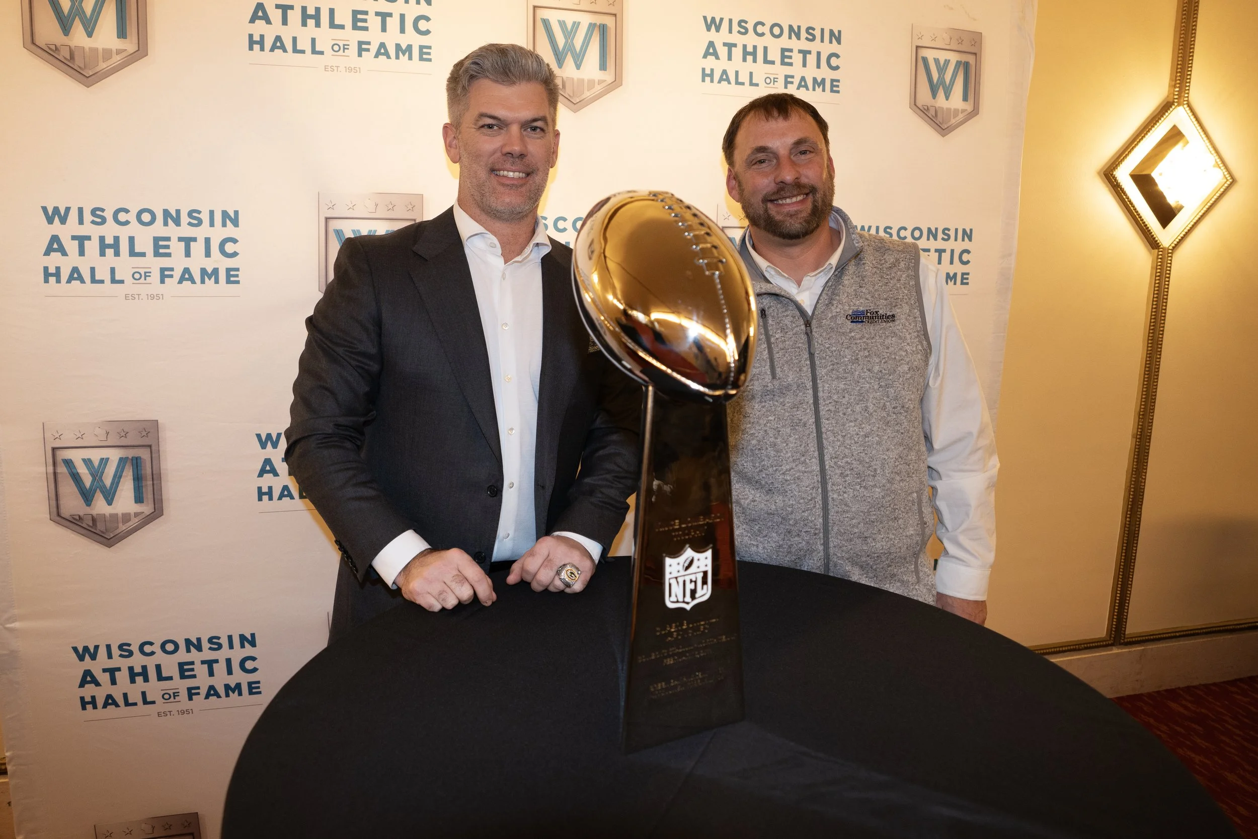 Two men standing behind a table with the NFL MVP trophy, at the Wisconsin Athletic Hall of Fame event.