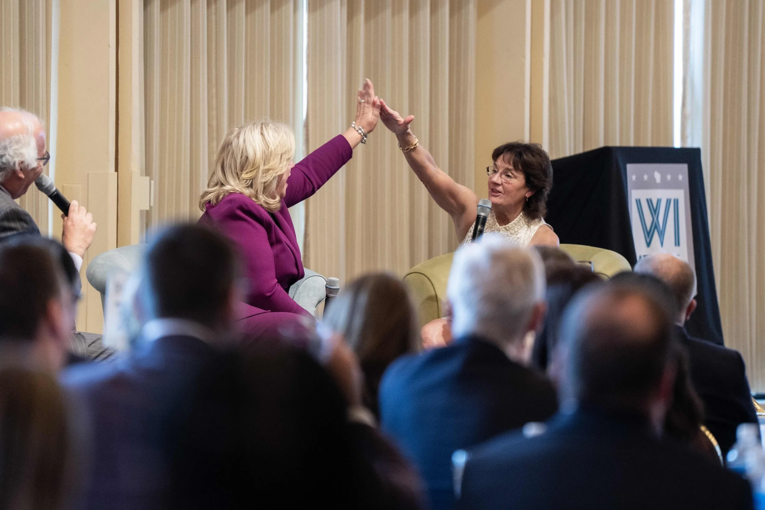 Two women giving each other a high five during a panel discussion at an event, with an audience listening.