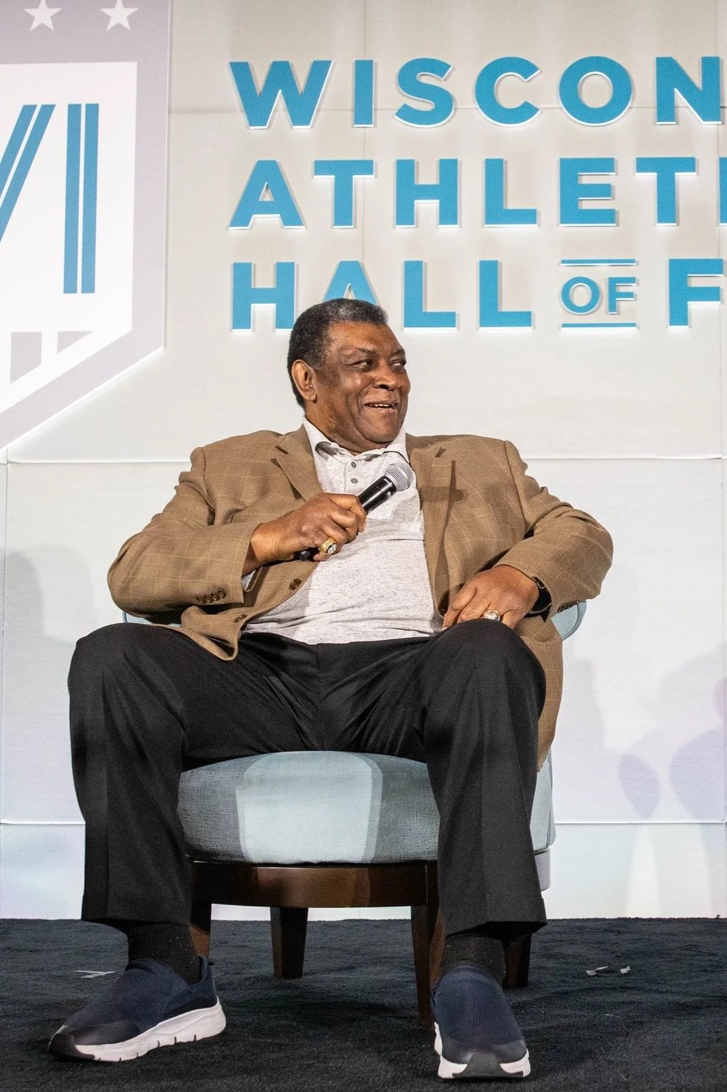 A man sitting on a chair holding a microphone, in front of a background with the text 'Wisconsin Athletic Hall of Fame' and a logo.
