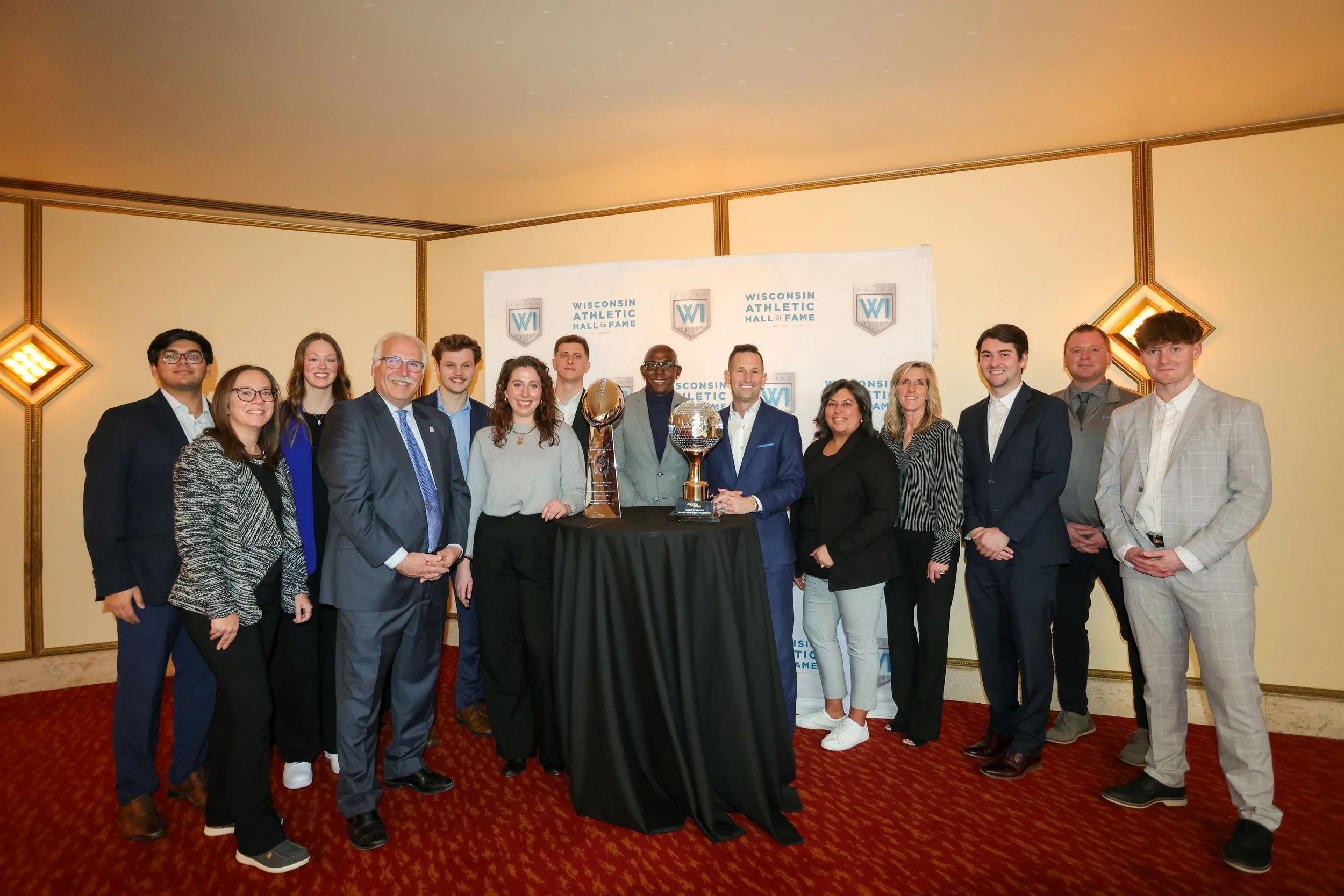 Group of people posing for a photo at the Wisconsin Athletic Hall of Fame awards ceremony, with trophies on a table in front of them.