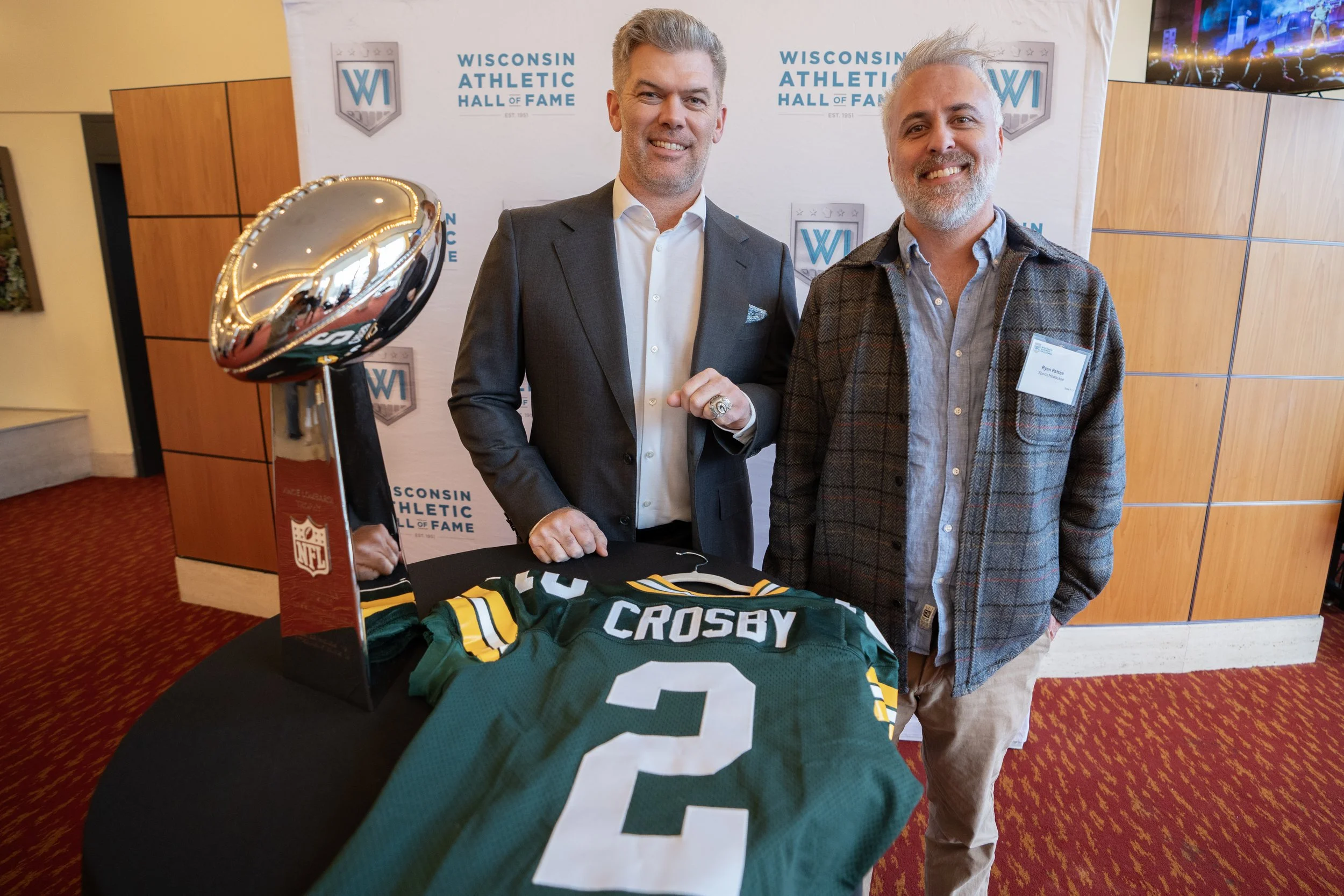 Two men stand behind a table with a Green Bay Packers football jersey labeled 'Crosby' and the number 2, and a large NFL trophy with a football on top. They are at the Wisconsin Athletic Hall of Fame event.