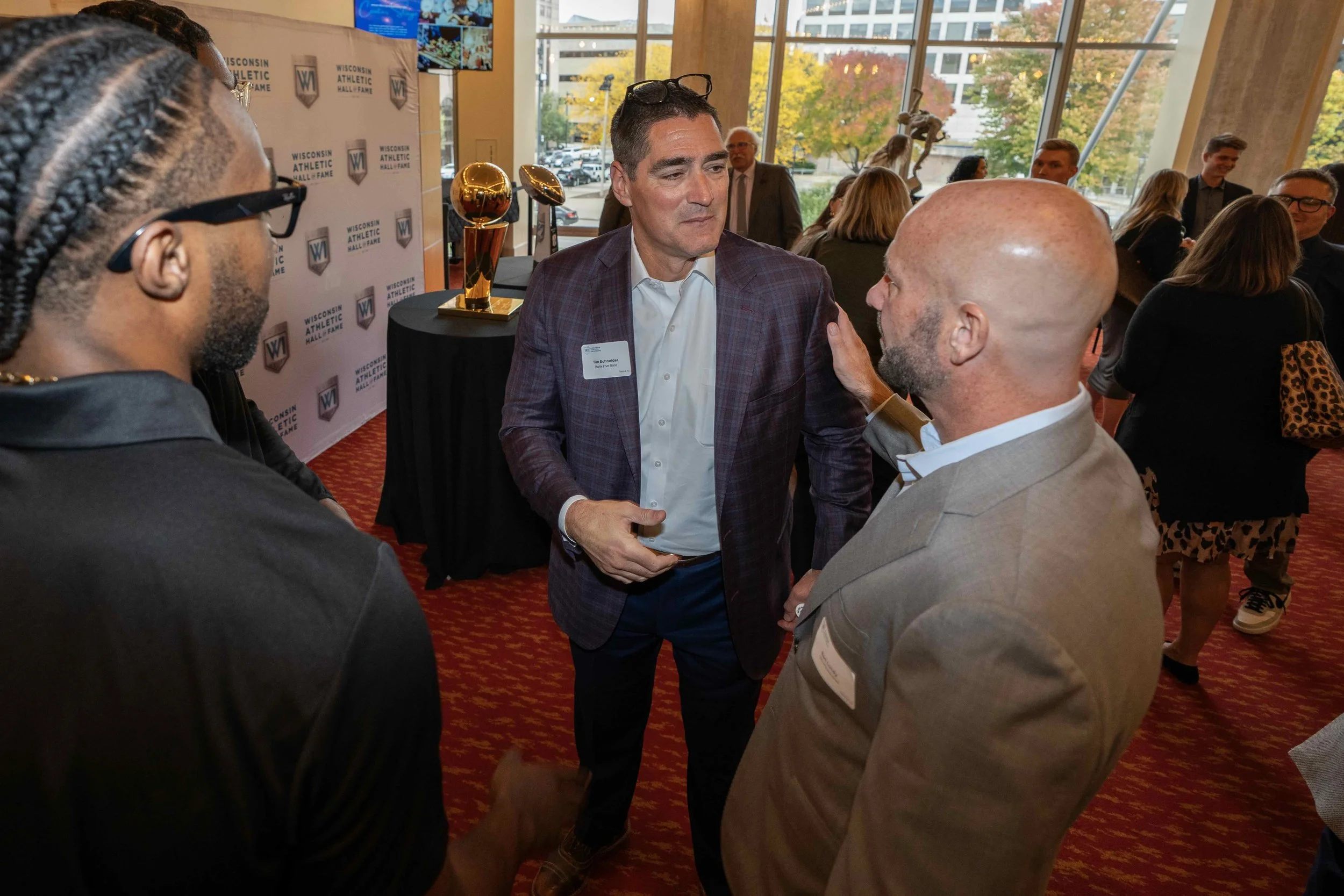 People at a networking event inside a modern building with large windows and fall foliage outside. Three men are in the foreground, one with a plaid blazer talking to another with a beard and tan blazer. In the background, others are mingling near a 