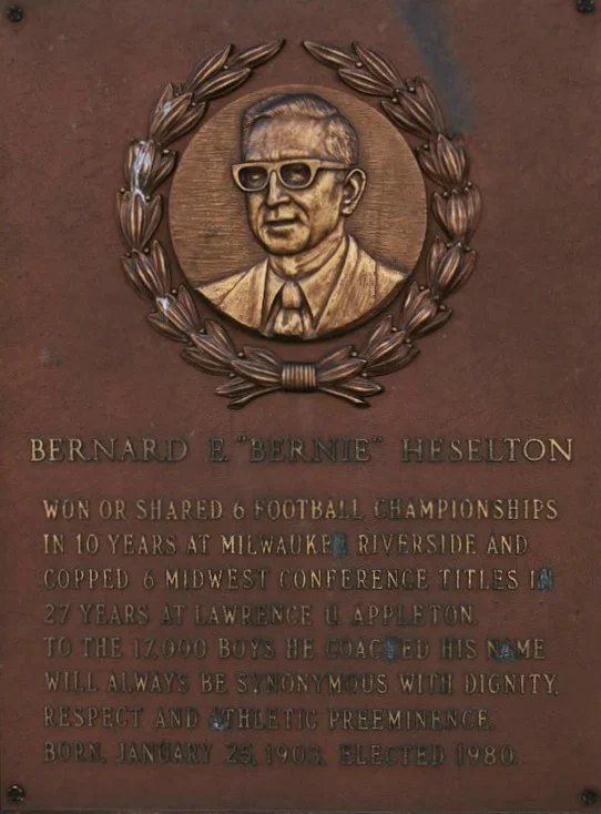 Plaque with a portrait of Bernard E. 'Bernie' Hesleton surrounded by a laurel wreath. The plaque contains text describing his achievements in football, coaching, and personal details, including his birth date and year elected.