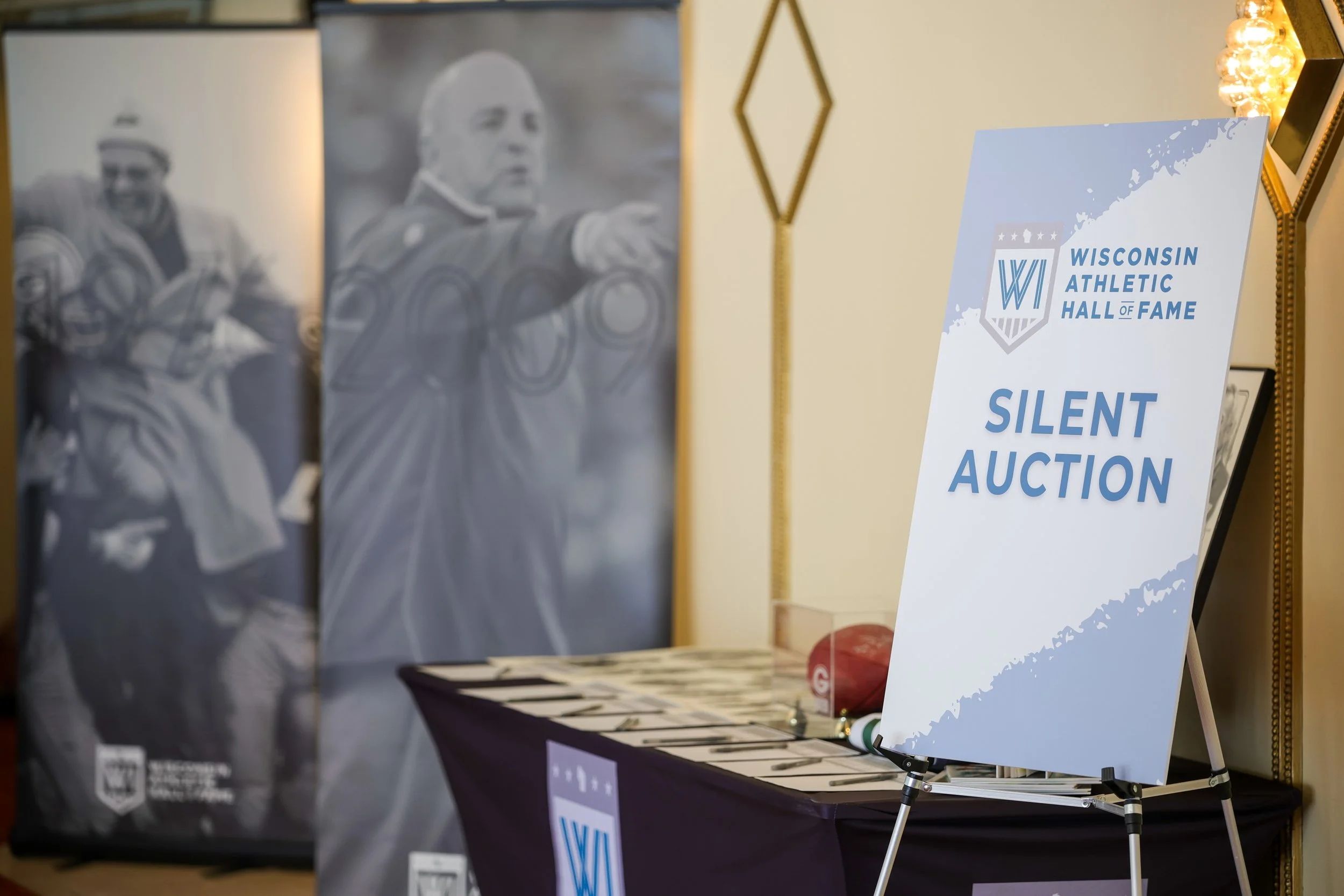 Sign for Wisconsin Athletic Hall of Fame Silent Auction next to table with auction items, background features black and white photos of athletes.