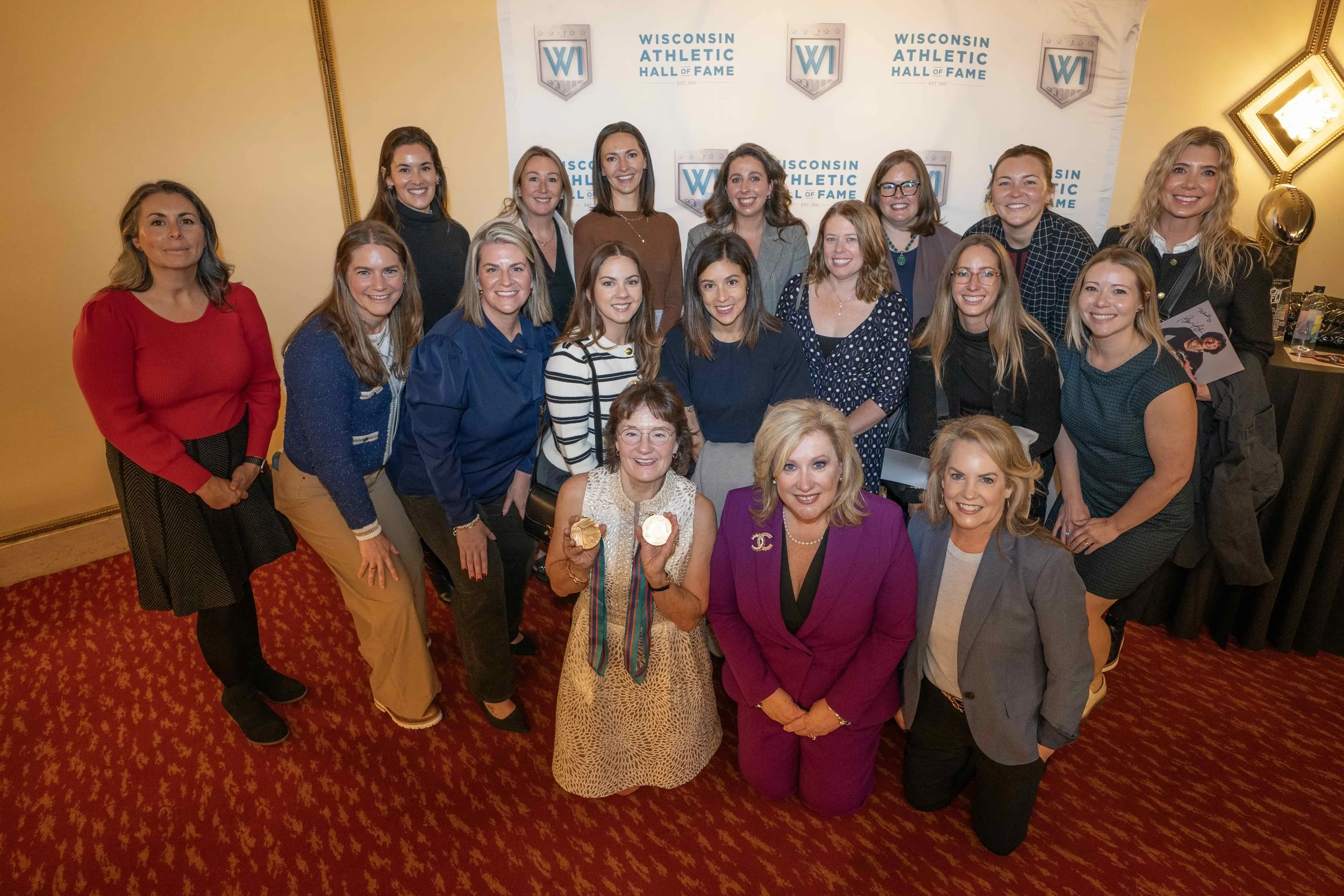 Group of women at a Wisconsin Athletic Hall of Fame event, posing together on a red carpet with a backdrop that displays the Wisconsin Athletic Hall of Fame logo.