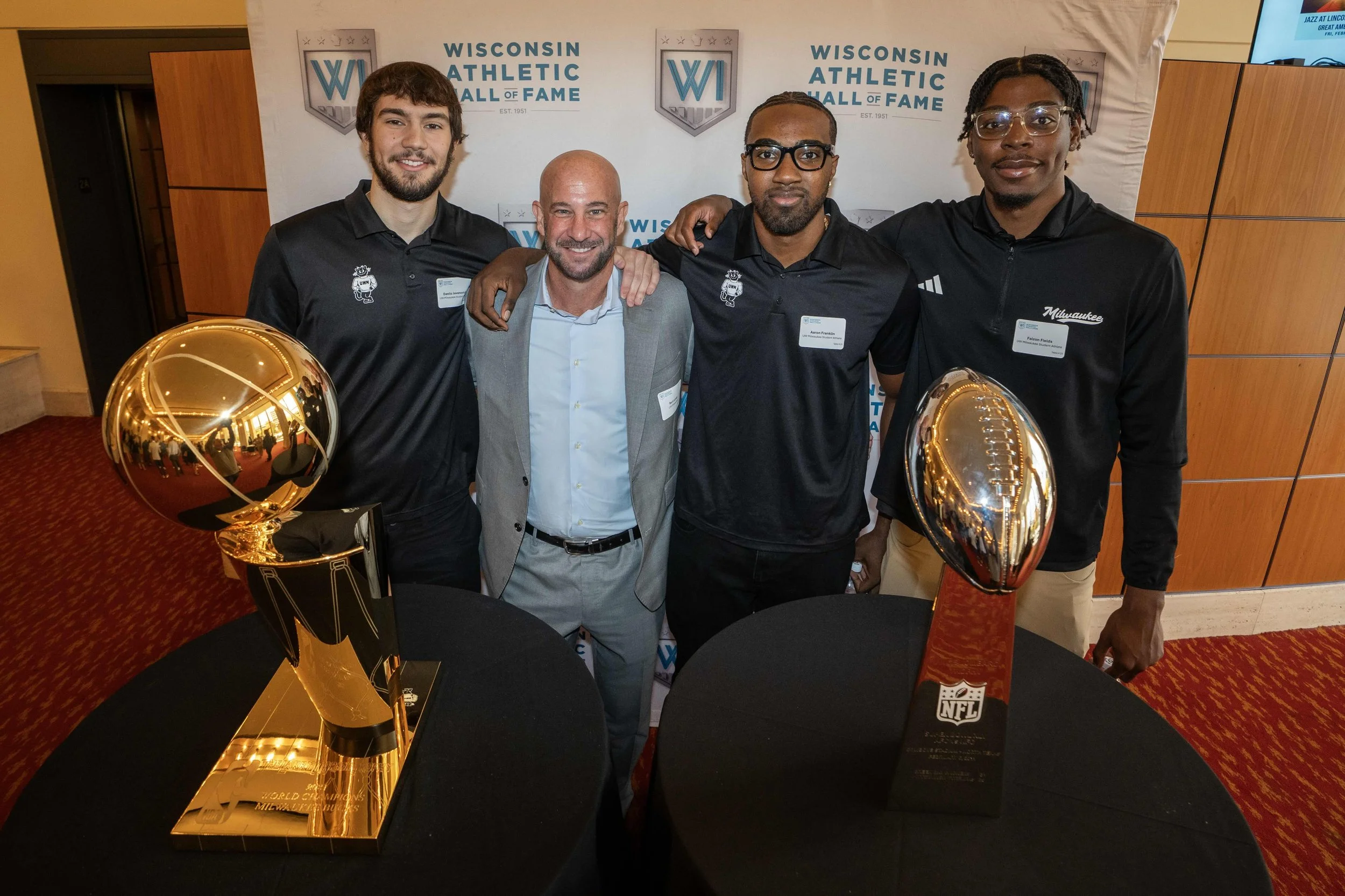 Four men posing for a photo at the Wisconsin Athletic Hall of Fame, with two Super Bowl trophies on tables in front of them.