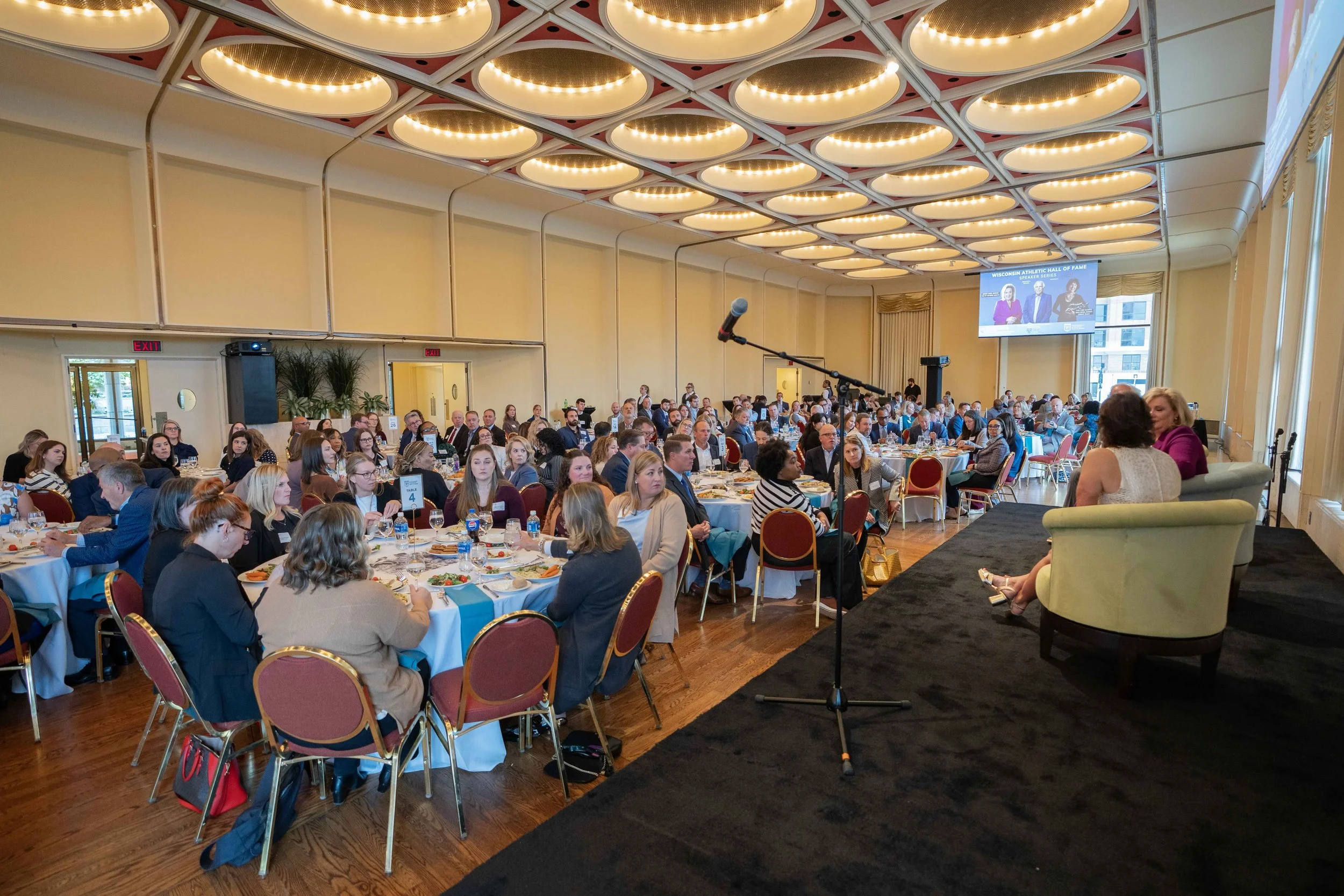 Large conference room filled with round tables of attendees listening to a speaker on stage. The stage has a small sofa and a woman speaking into a microphone, with a large screen showing a presentation behind her. The room has a high ceiling with ci