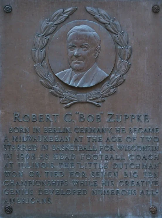 A memorial plaque with a portrait of Robert C. 'Bob' Zuppke. The plaque includes a biography stating he was born in Berlin, Germany, became a Milwaukeean at age two, starred in basketball for Wisconsin, and was a head football coach at Illinois. It mentions his seven Big Ten championships and his contribution to American sports.