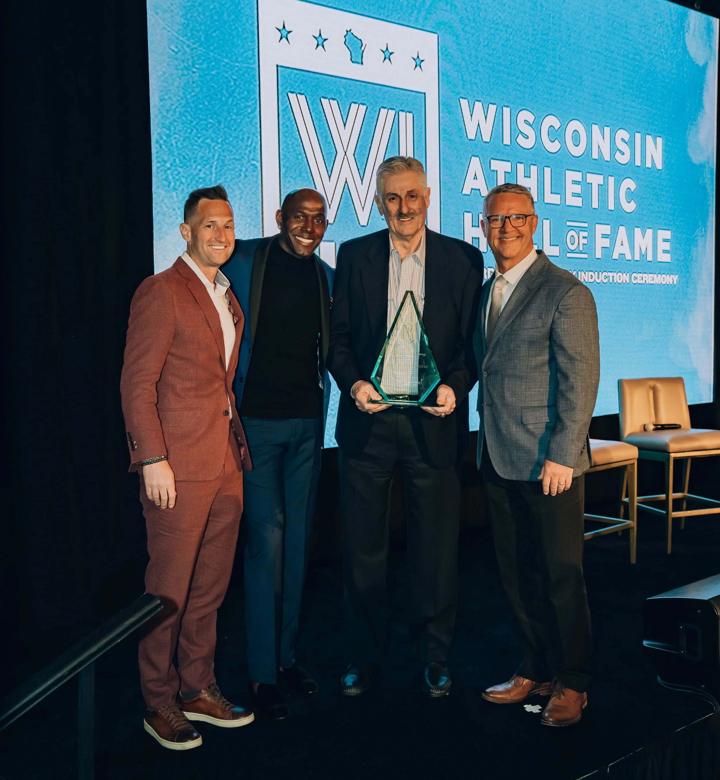 Four men are standing on a stage at an event, with a large digital screen behind them displaying the Wisconsin Athletic Hall of Fame logo and text. They are dressed in formal attire, smiling, and one of them is holding an award.