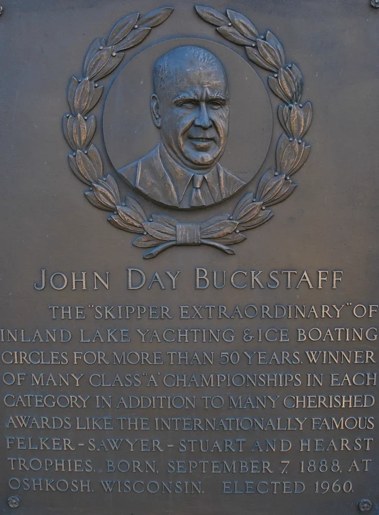 A bronze plaque with a portrait of John Day Buckstaff wearing a suit and tie, surrounded by a laurel wreath. The plaque contains text describing his achievements in inland lake yachting and ice boating, his awards, and personal details including his birthdate, birthplace, and year of election.